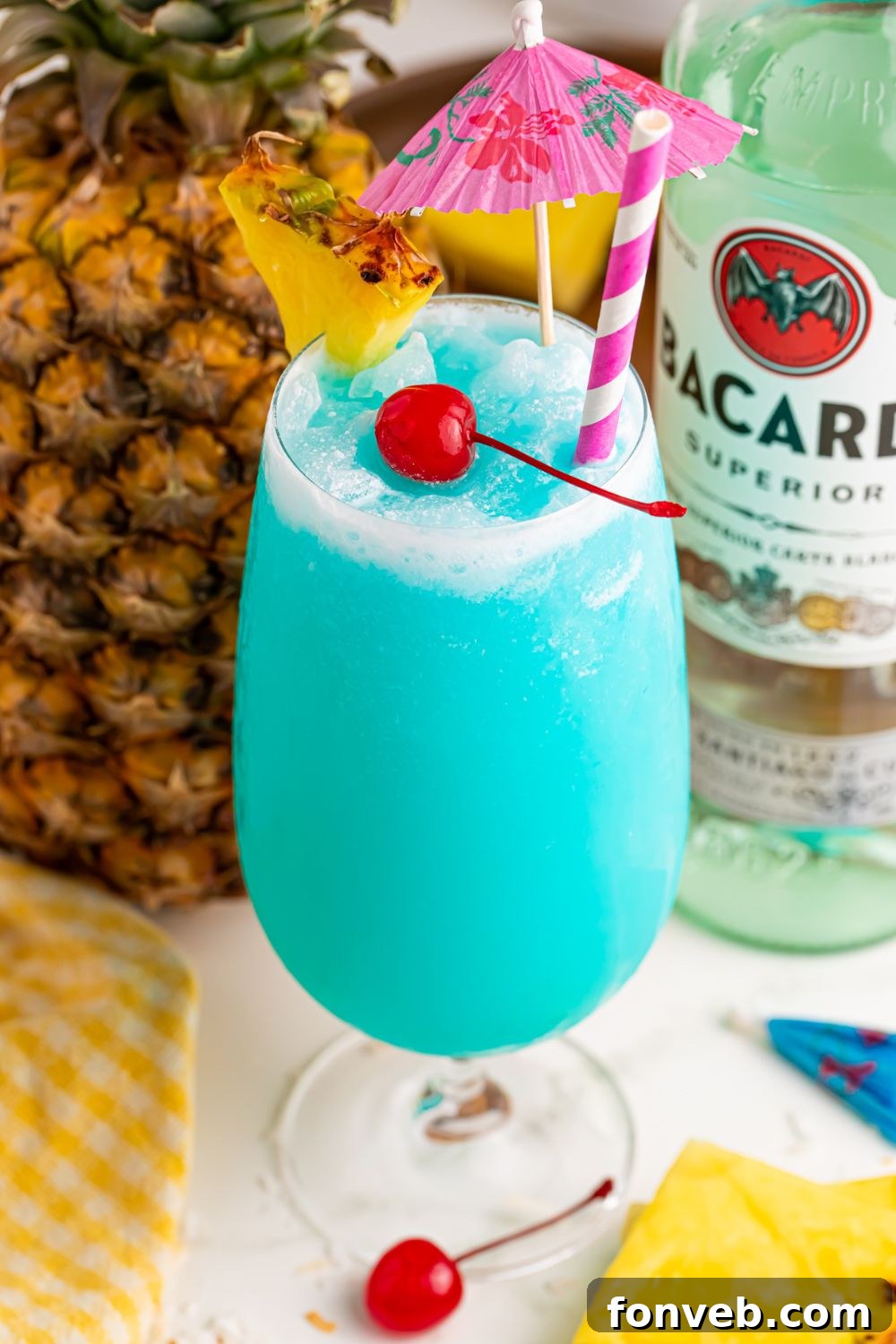 pineapple and bacardi rum on table with a glass of Frozen Blue Hawaiian Cocktail in it and a cherry and pineapple wedge in front of it 