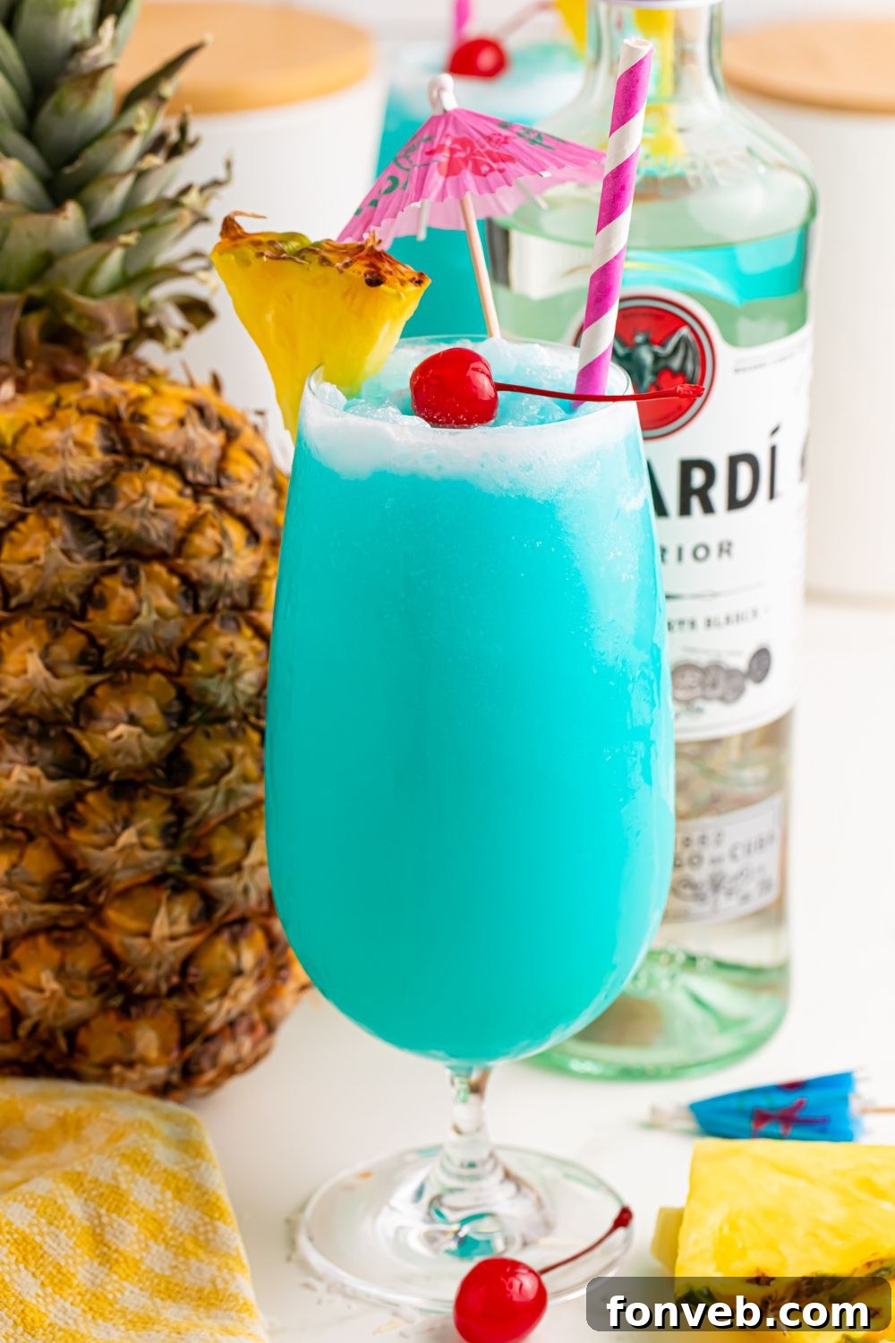 view of Frozen Blue Hawaiian Cocktail with umbrella and straw in glass sitting on a table with ingredients used in it scattered on table 