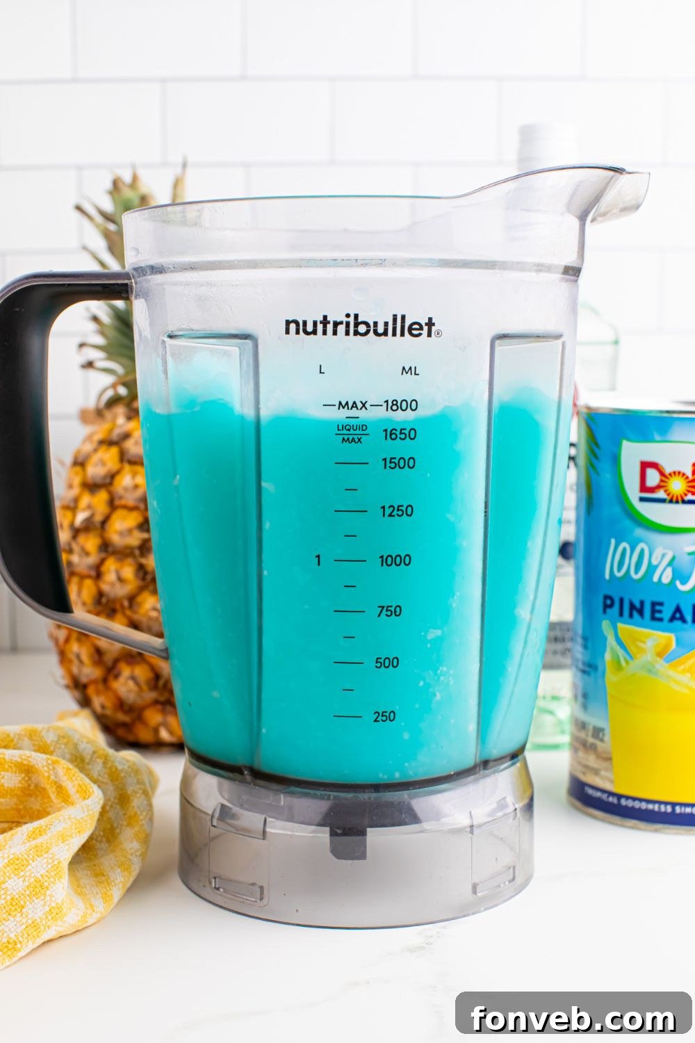 side view of Frozen Blue Hawaiian Cocktail in a blender on table with pineapple juice and whole pineapple behind it on table