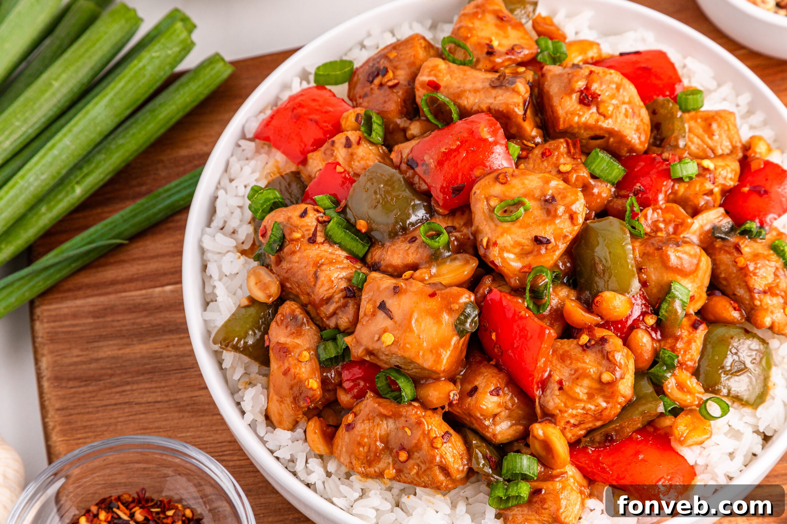 A bowl of Better Than Takeout Kung Pao Chicken on a bed of rice, with a cutting board of fresh vegetables nearby, ready for serving.