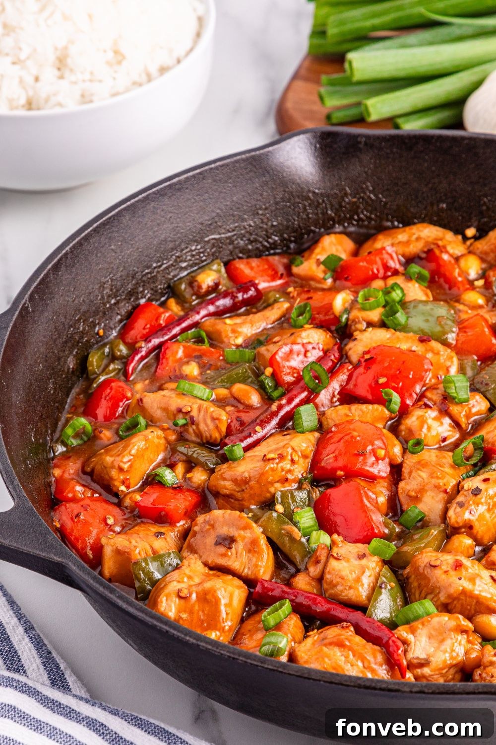 Better Than Takeout Kung Pao Chicken in a cast iron skillet, with a bowl of rice and fresh green onions on a cutting board, ready for serving.