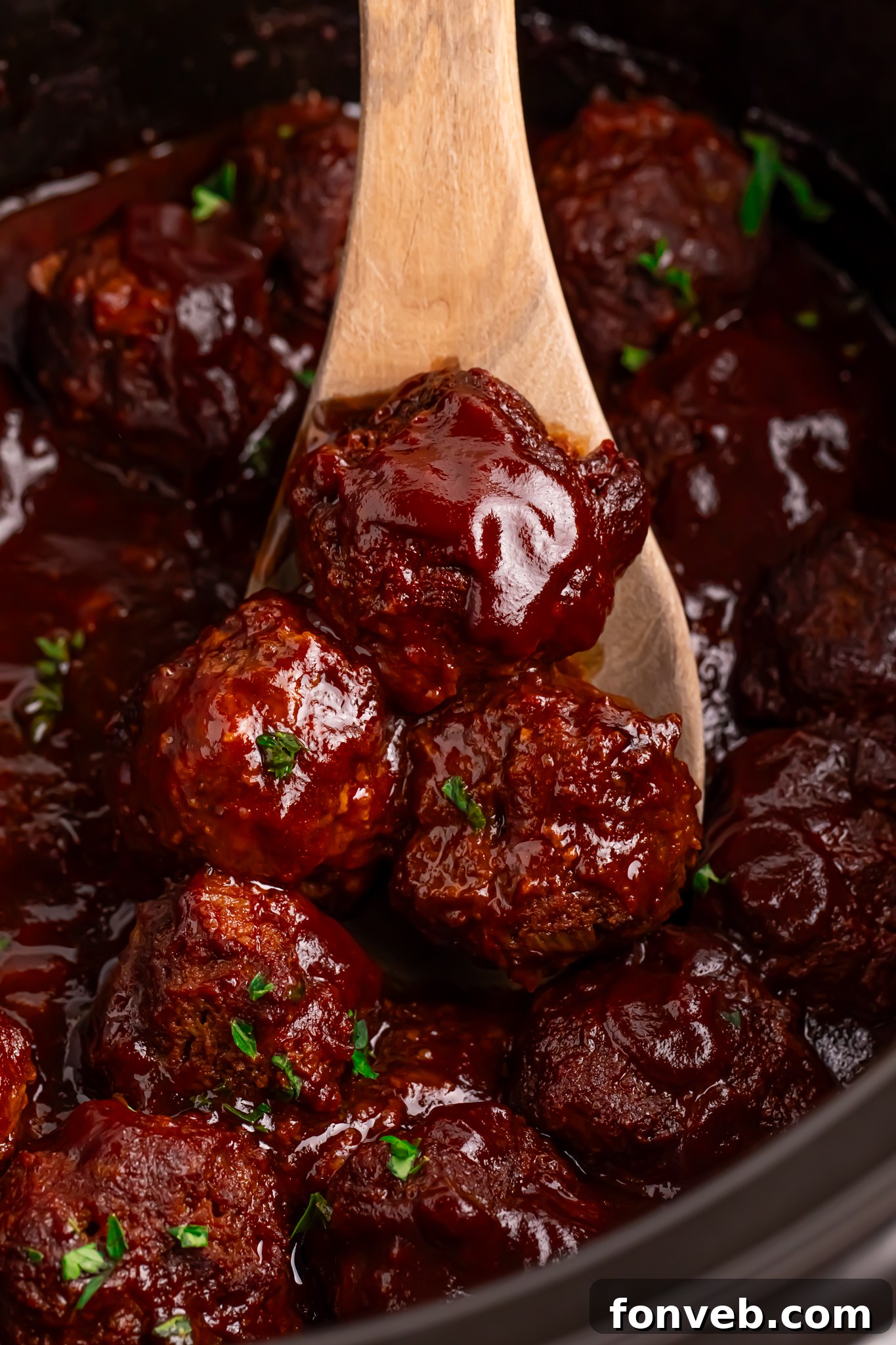 Crockpot Homemade BBQ Meatballs in a black slow cooker with a wooden spoon holding 3 up 