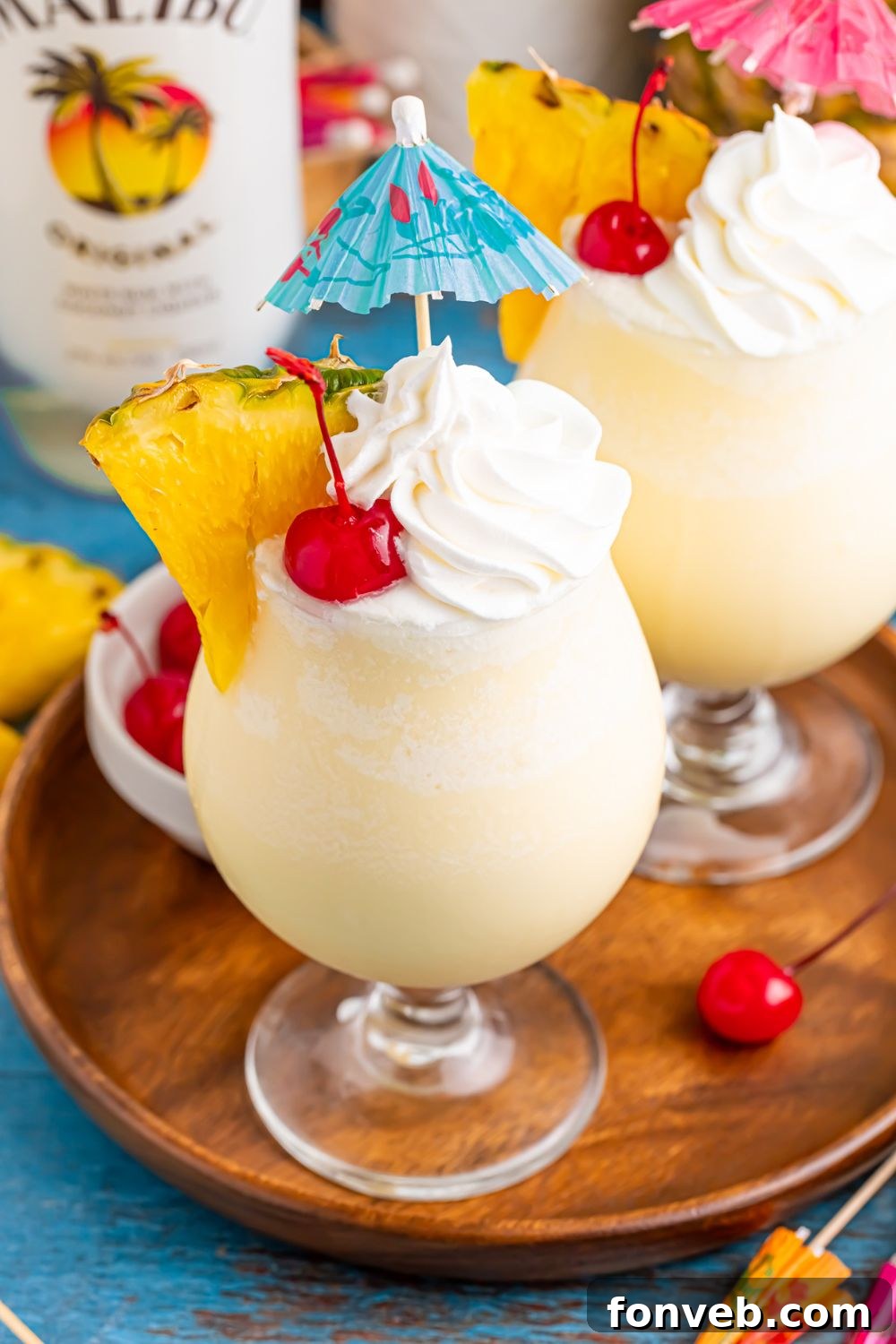 Creamy Frozen Malibu Piña Colada Delight 15 Frozen Malibu Piña Colada on table with pineapple wedges and whipped cream and umbrella topper
