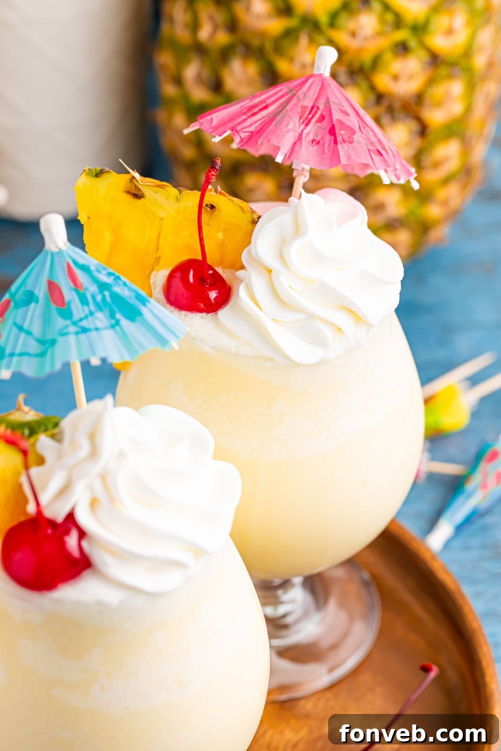 Creamy Frozen Malibu Piña Colada Delight 16 Frozen Malibu Piña Coladas on table with whipped cream, cherries, and umbrella, and more umbrella scattered on table