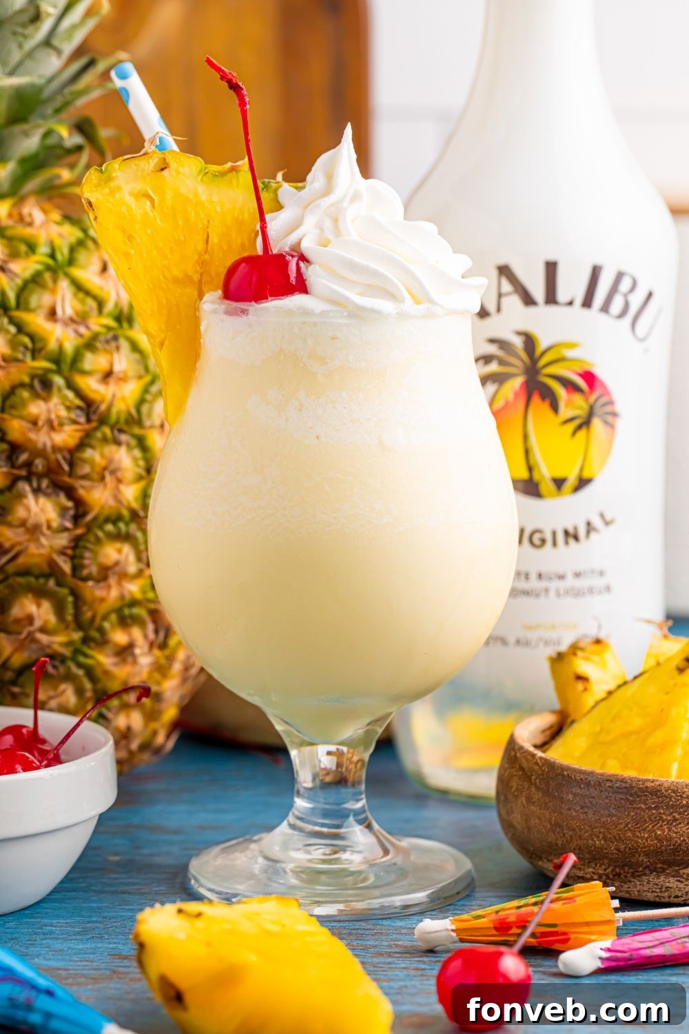 Creamy Frozen Malibu Piña Colada Delight 20 Frozen Malibu Piña Colada in a glass on table with pineapple slices and malibu and a whole pineapple to side on table