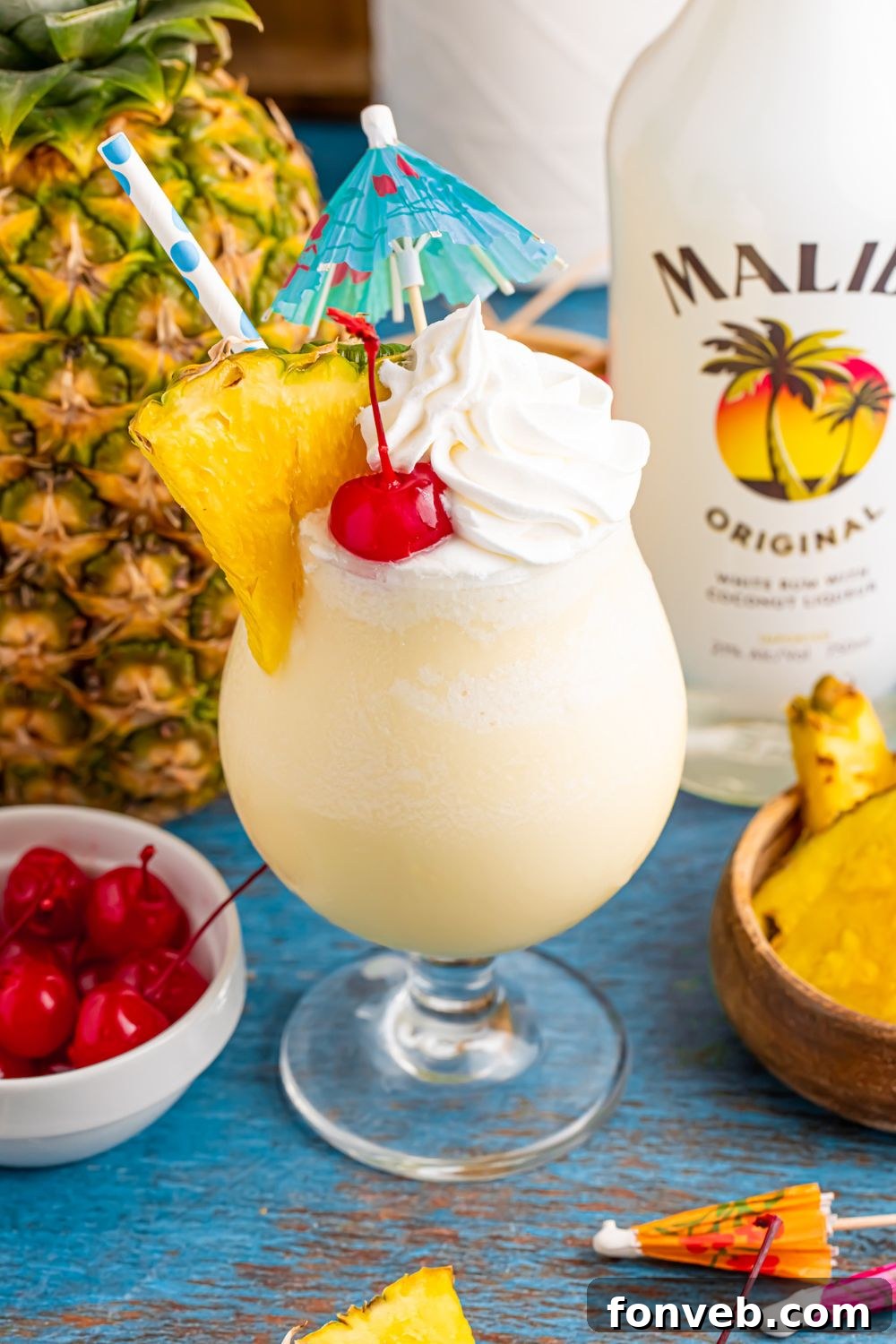 Creamy Frozen Malibu Piña Colada Delight 22 Frozen Malibu Piña Colada in a cocktail glass with a whole pineapple to side, bowl of cherries, and bottle of Malibu rum