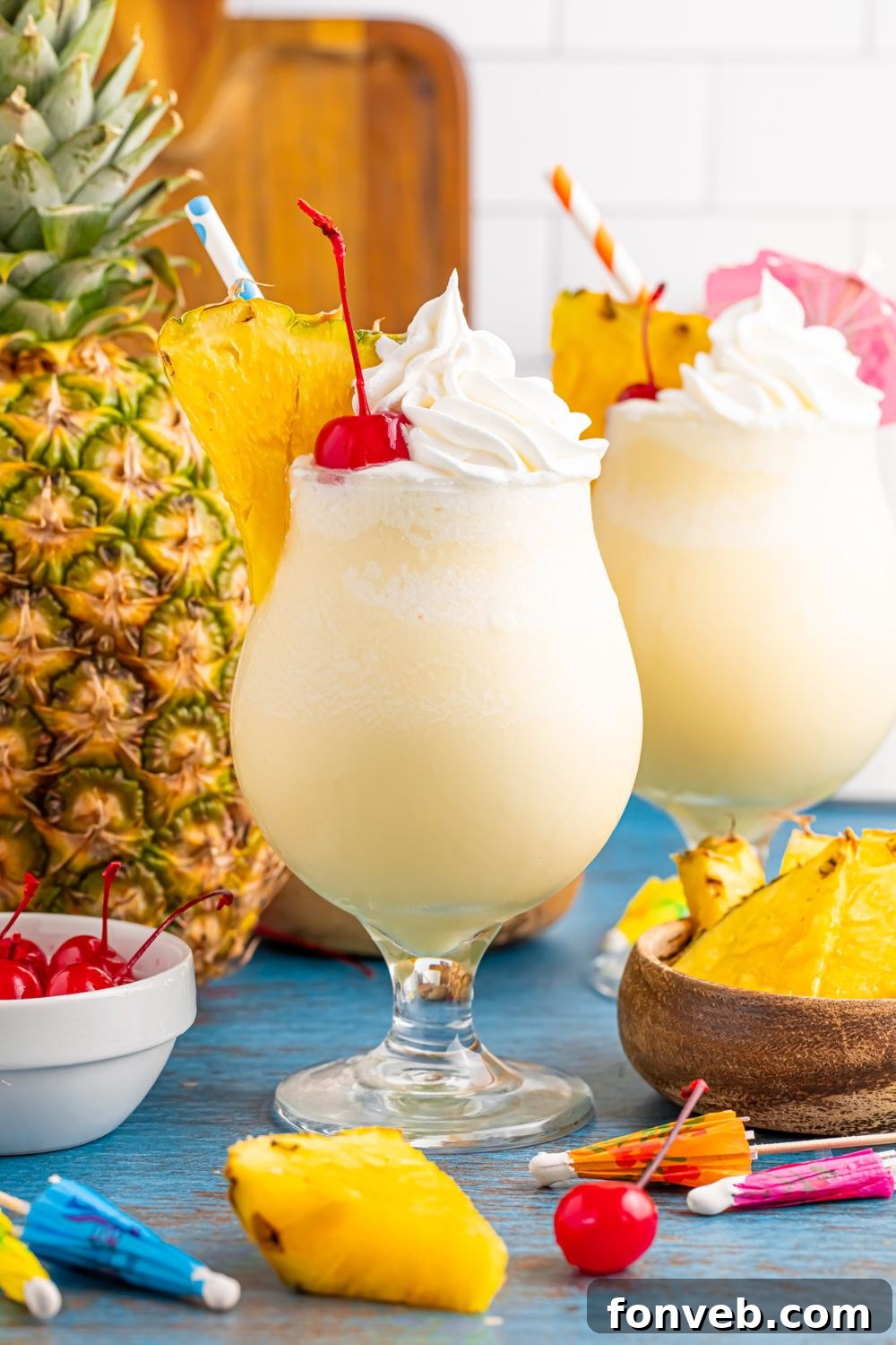 Creamy Frozen Malibu Piña Colada Delight 4 Frozen Malibu Piña Coladas on table with whole pineapple and cherries on table and umbrellas for drinks