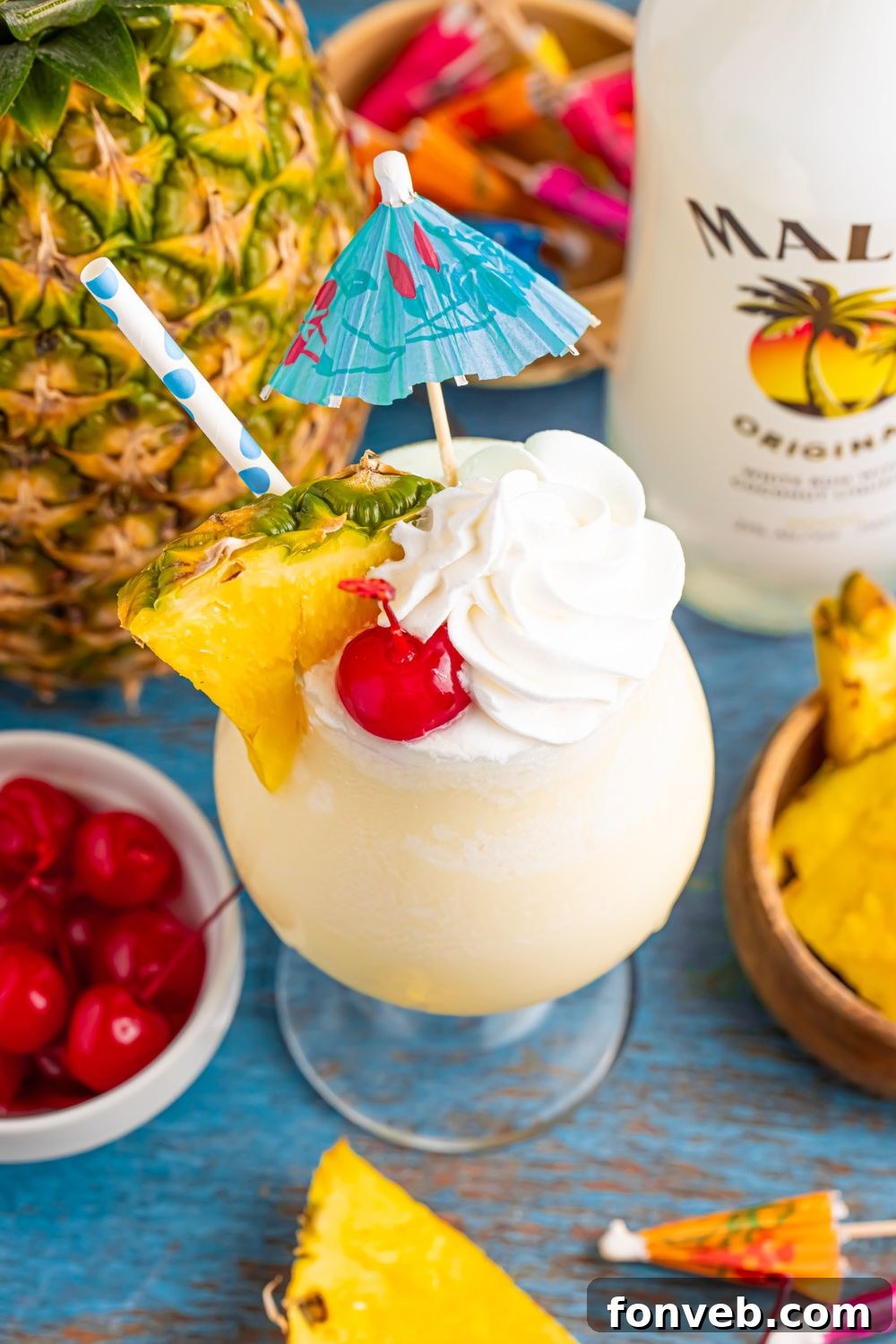 Creamy Frozen Malibu Piña Colada Delight 6 Frozen Malibu Piña Colada on table with a pineapple in the background and a bowl of cherries to side, and bottle of Malibu rum