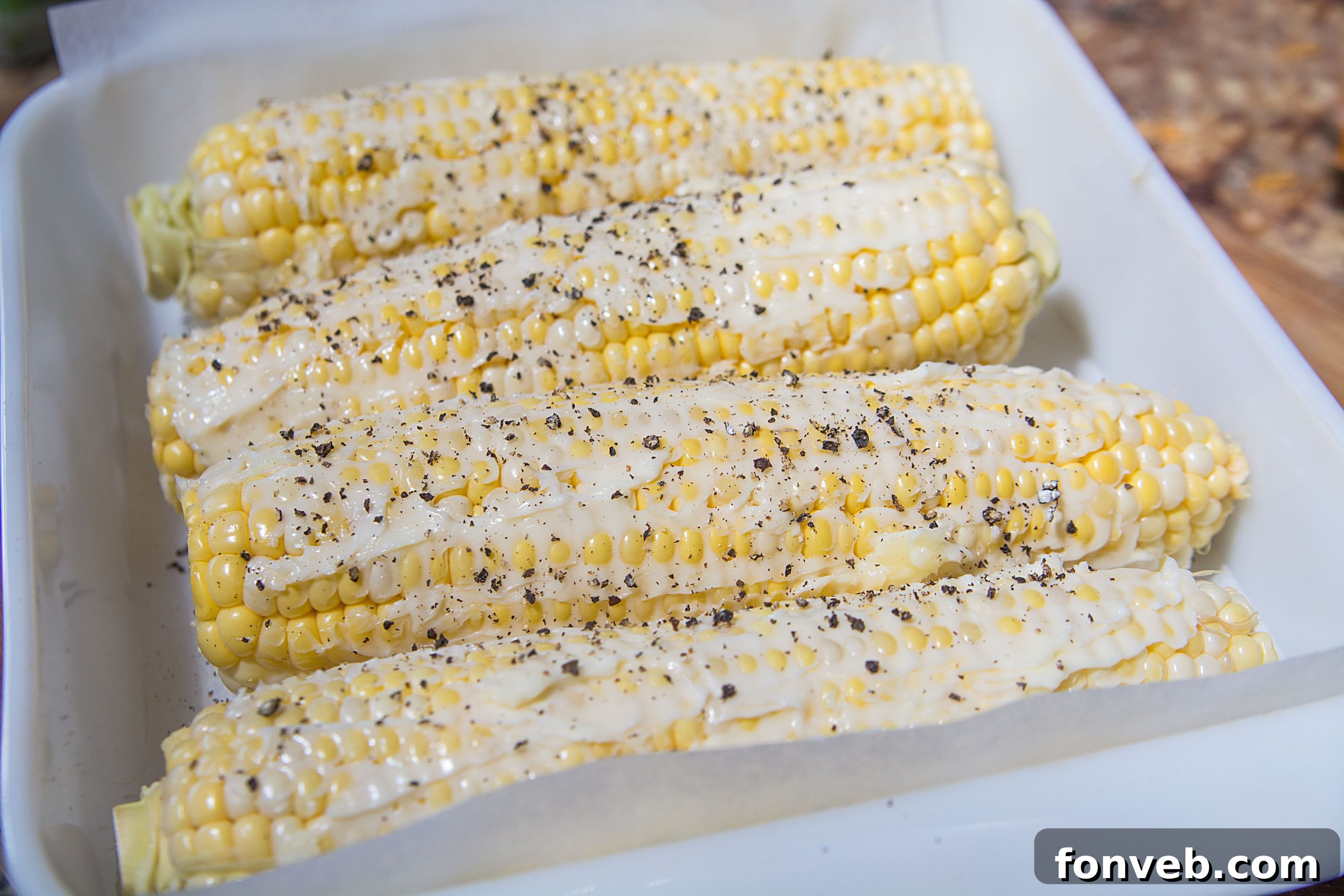 Close-up of a hand spreading butter and spices on an ear of corn before roasting, showcasing the preparation process.