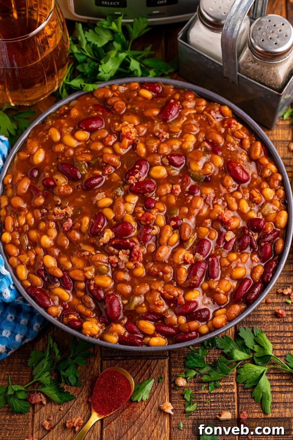 Crockpot bourbon baked beans in a bowl on table with some fresh herbs scattered on table and a towel 