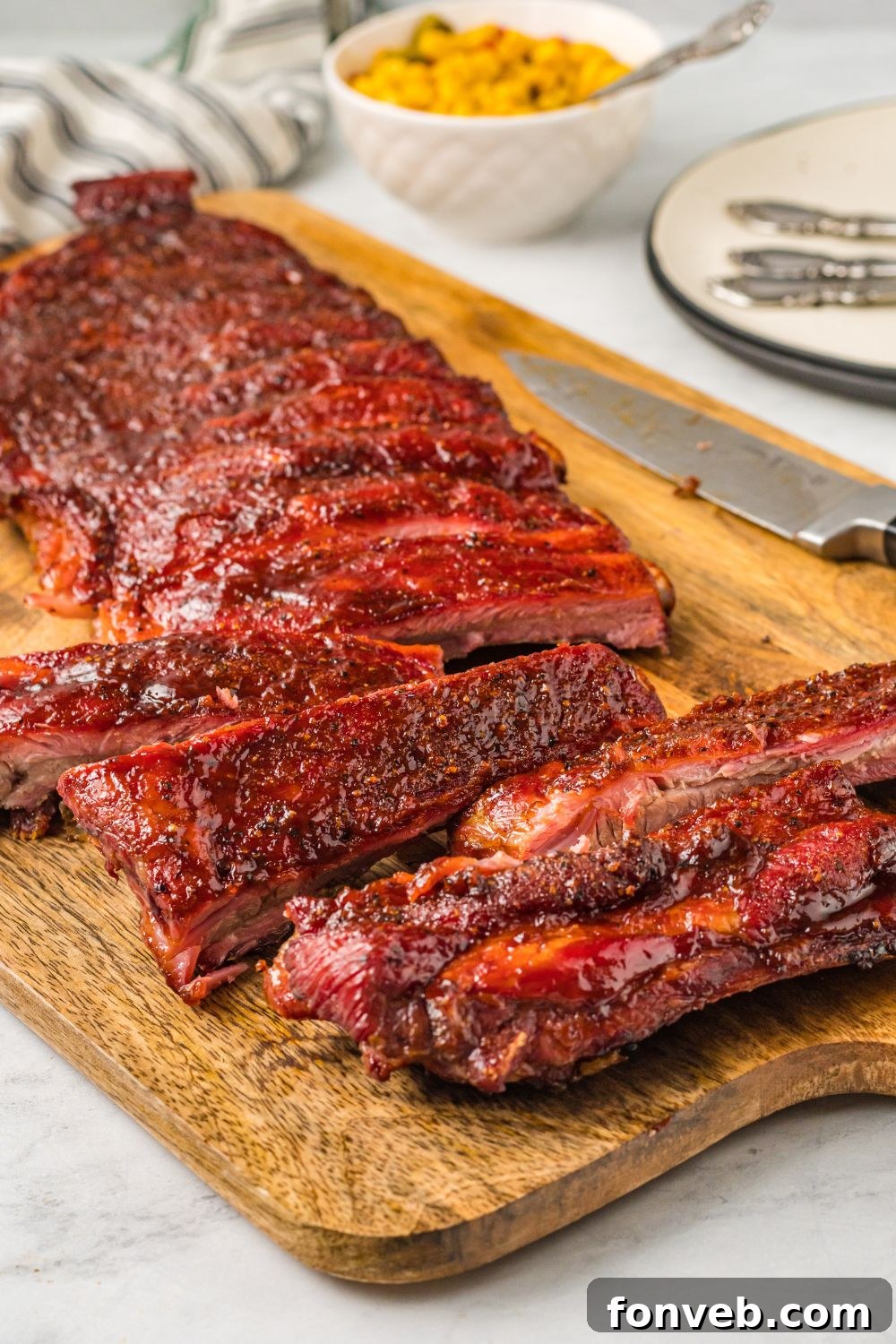 Grilled BBQ Baby Back Ribs sliced up on a cutting board with a bowl of corn in background