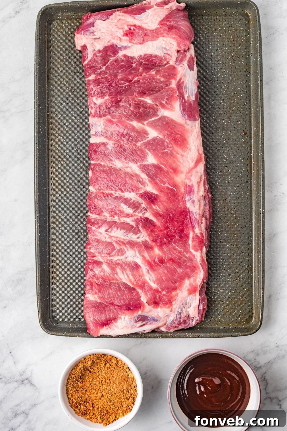 raw ribs on a cooking tray with bbq sauce and dry rub in a bowl