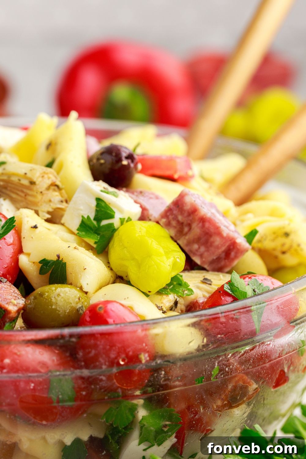 side view of Antipasto Tortellini Pasta Salad in a glass bowl with spoons pushed into bowl and vegetables around the table 