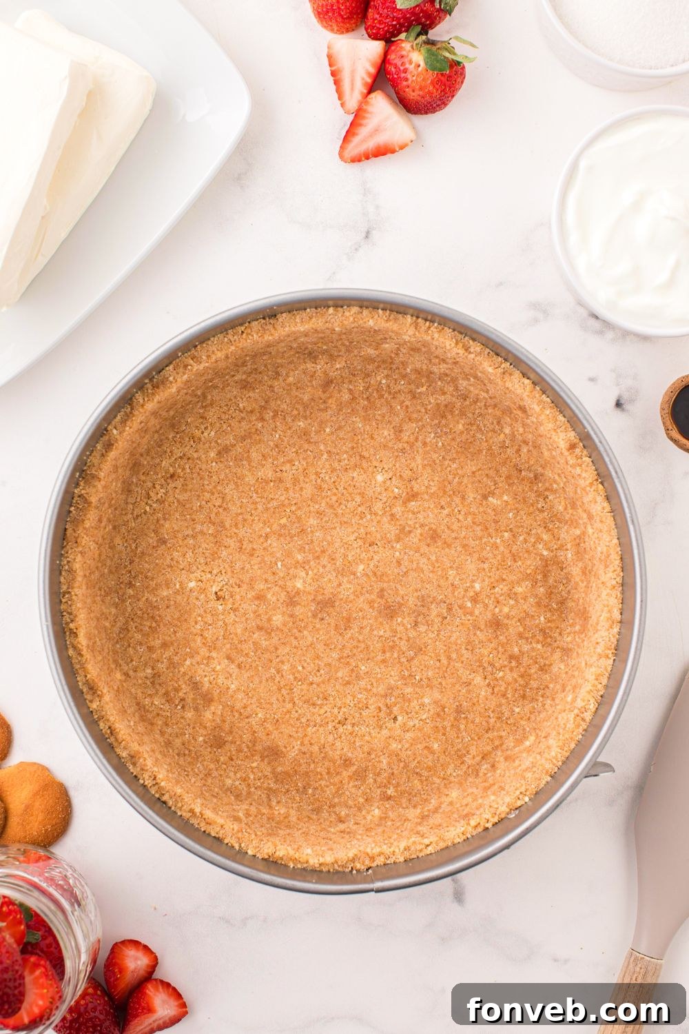 vanilla wafer crust in a pie plate on table with strawberries and other ingredients around it for making a No Bake Strawberry Cheesecake