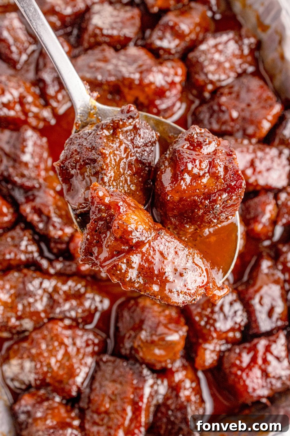 A scoop of luscious Chuck Roast Burnt Ends is lifted from a foil pan with a spoon, showcasing the tender meat and rich, sticky sauce, ready to be enjoyed.
