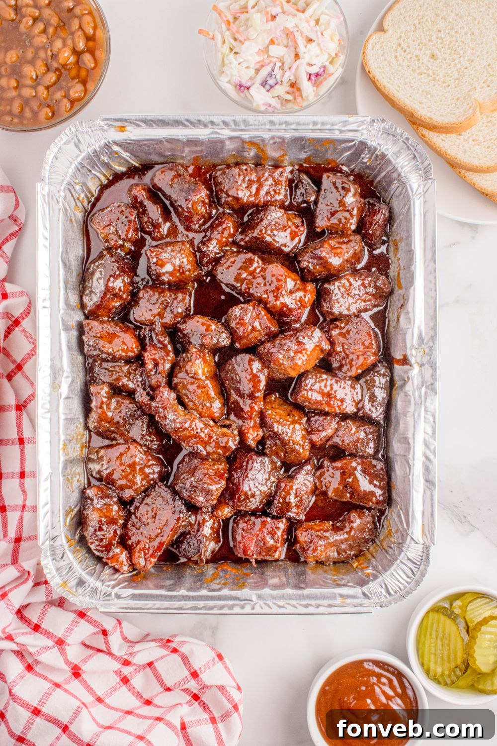 Chuck Roast Burnt Ends are served family-style in a large tin foil pan, surrounded by slices of fresh bread, crisp coleslaw, and tangy pickles, making for a perfect BBQ spread.