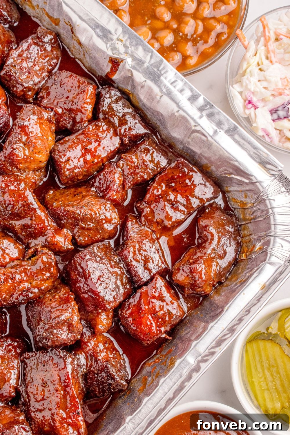Chuck Roast Burnt Ends are beautifully displayed in a large foil pan on a table, accompanied by bowls of hearty baked beans and creamy coleslaw, forming a delightful BBQ spread.