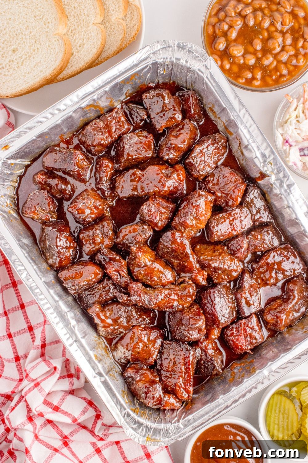 Chuck Roast Burnt Ends are arranged enticingly in a foil baking pan on a table, accompanied by slices of fresh bread, a bowl of savory beans, a bottle of ketchup, and a dish of crisp pickles, inviting a feast.