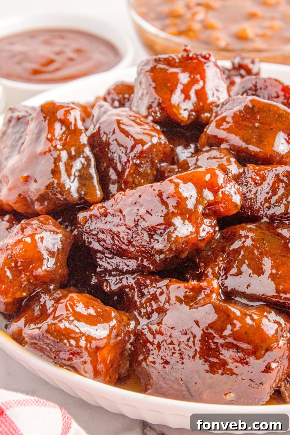 Chuck Roast Burnt Ends are generously piled in a white bowl on a table, with a small bowl of barbecue sauce placed to the side, ready for dipping.