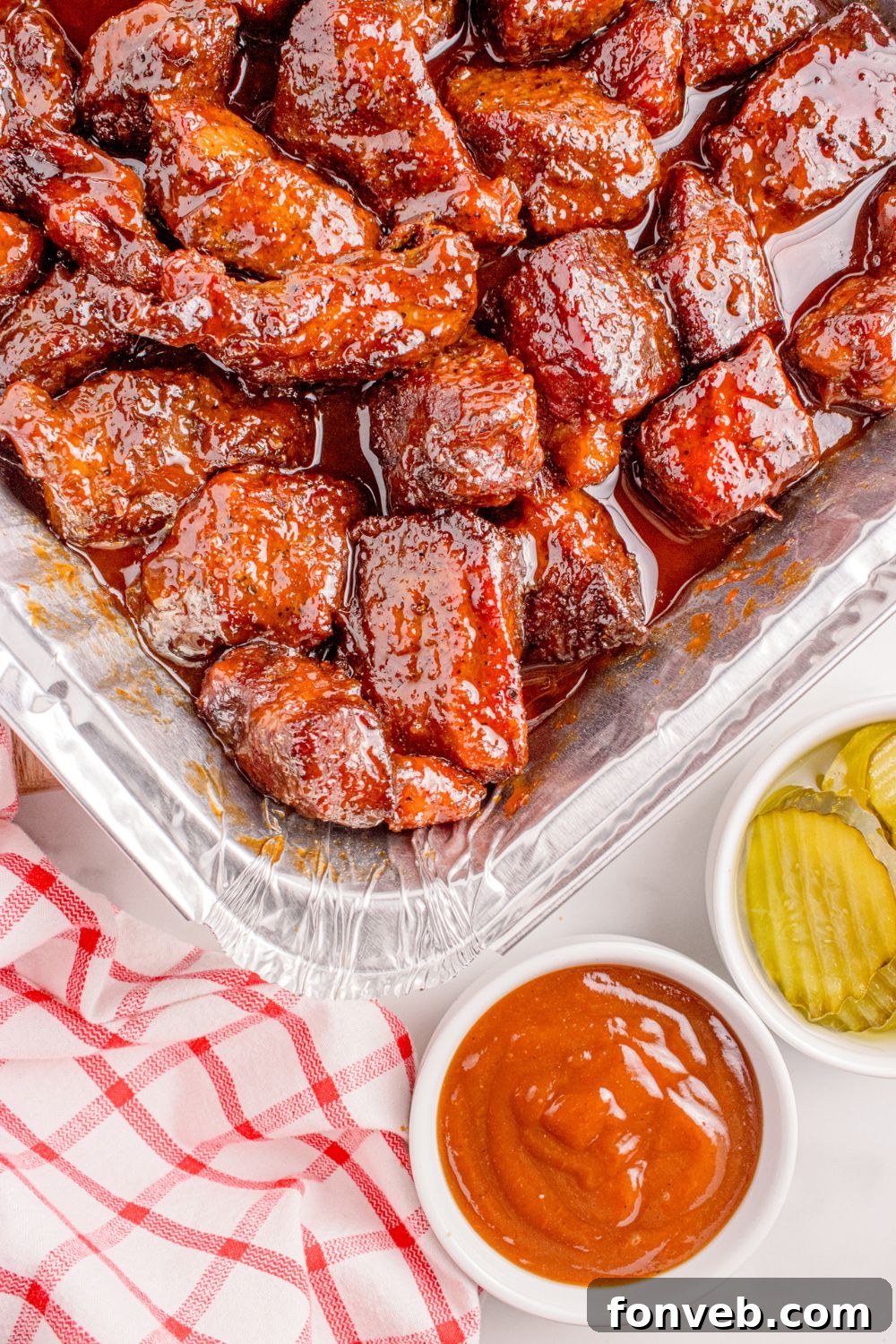 Smoked Chuck Roast Burnt Ends are presented in a rustic foil tray, ready for serving. Adjacent to the tray, small bowls hold an array of complementary sauces and crisp pickles, enhancing the BBQ experience.