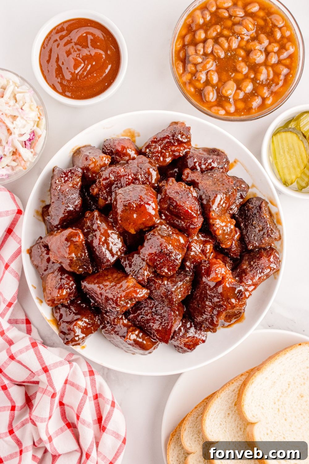 A heaping portion of Chuck Roast Burnt Ends is served in a bowl, flanked by a side dish of savory beans, crisp pickles, and a generous drizzle of barbecue sauce, ready for a delicious meal.