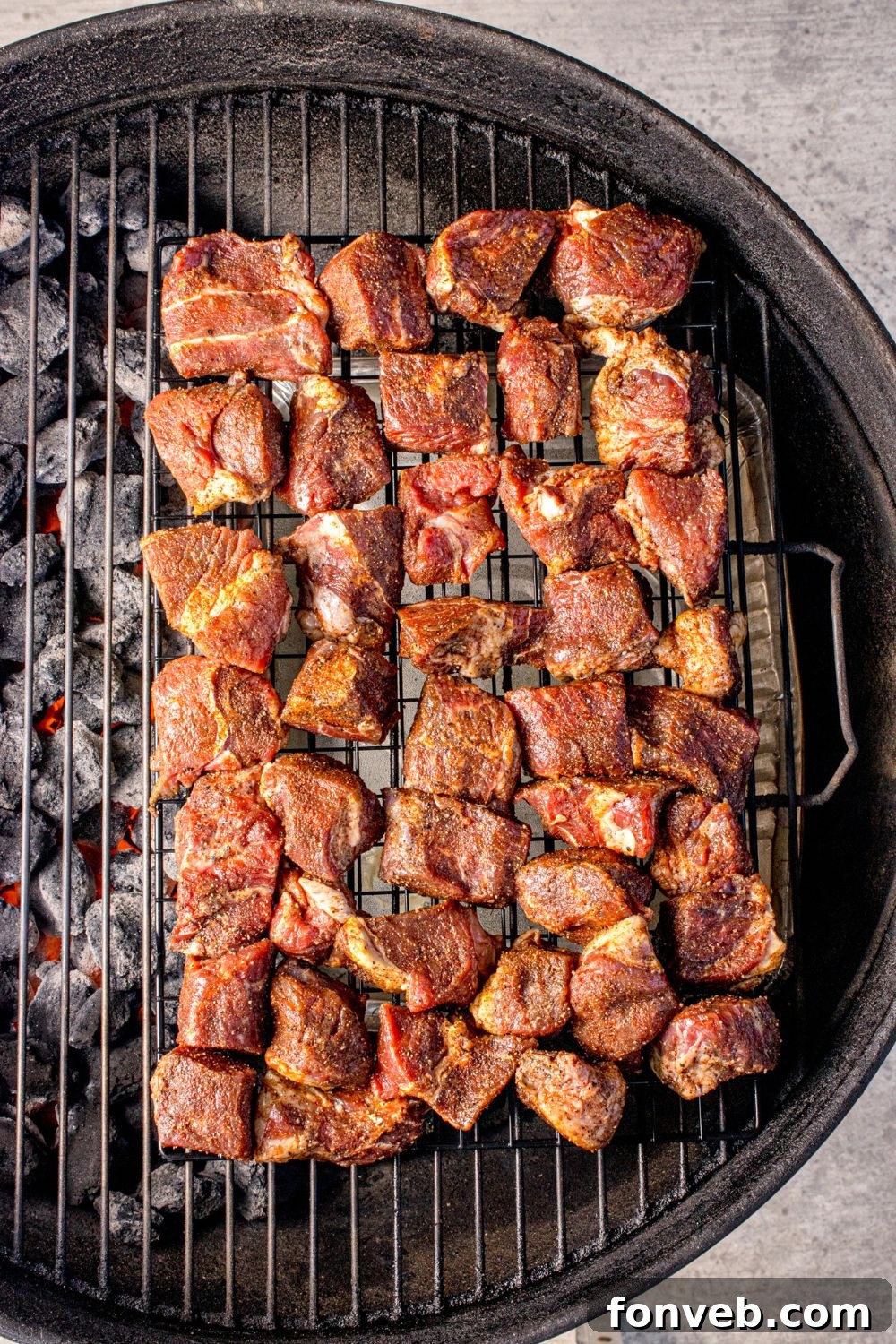 A charcoal grill, meticulously prepared for smoking, features pieces of seasoned steak spread evenly across the grates, absorbing the smoky essence.