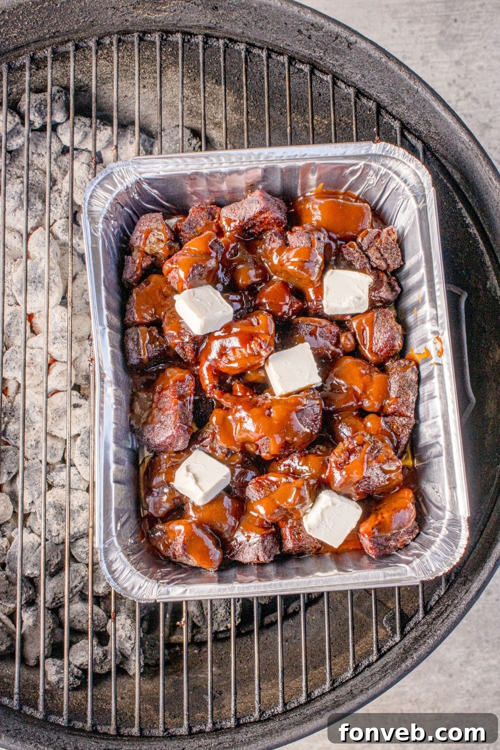 Chuck Roast Burnt Ends are simmering in a foil pan on a charcoal grill, with cubes of butter melting into the rich BBQ sauce, creating a luscious glaze.