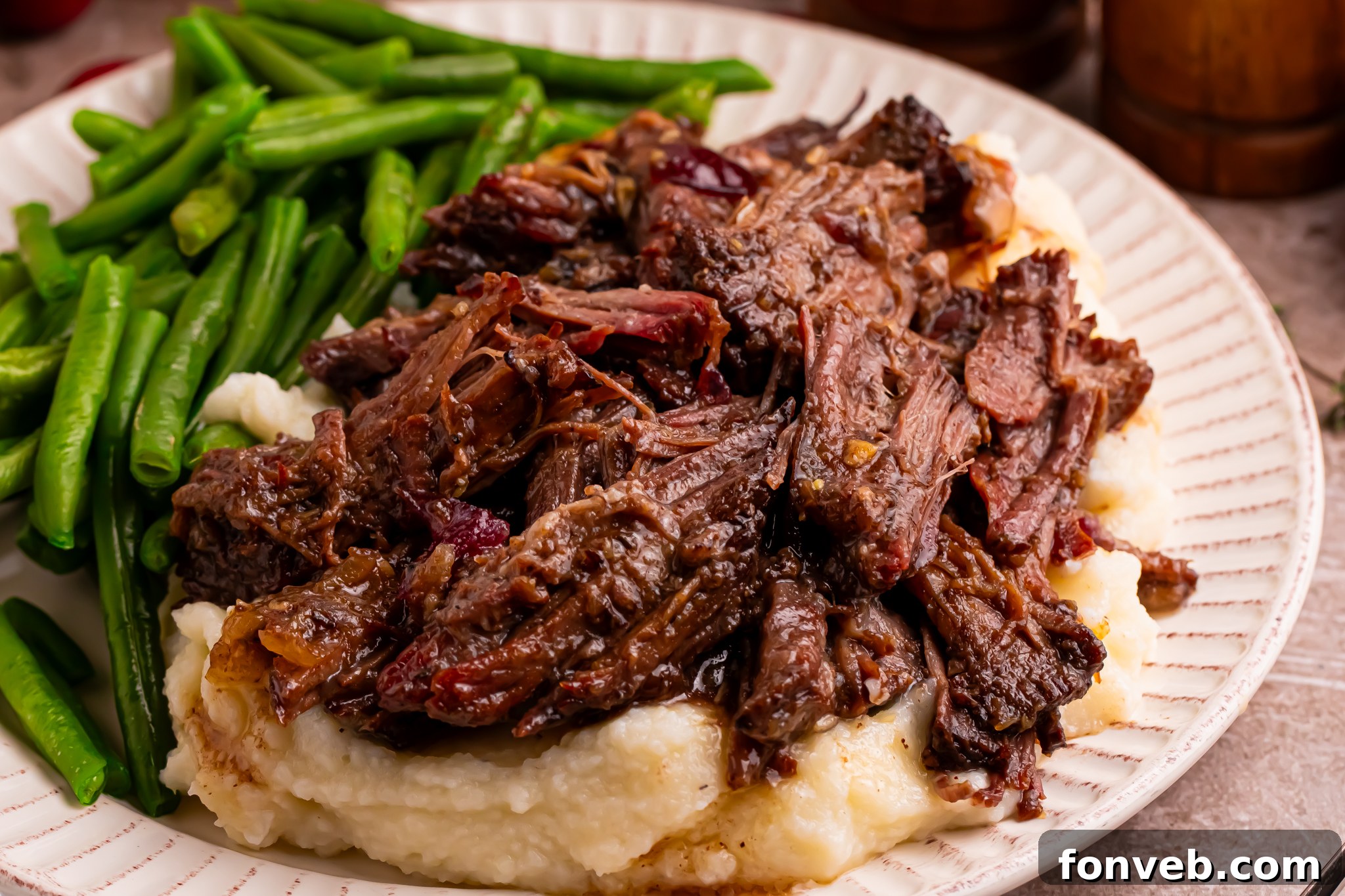Juicy Cranberry Pot Roast shredded on a white plate with mashed potatoes under and a side of green beans 