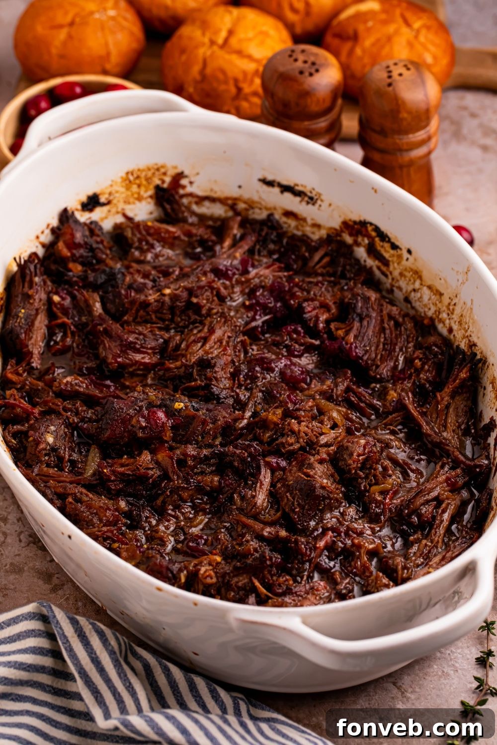 shredded cranberry pot roast in a casserole pan on table with dinner rolls behind it on table