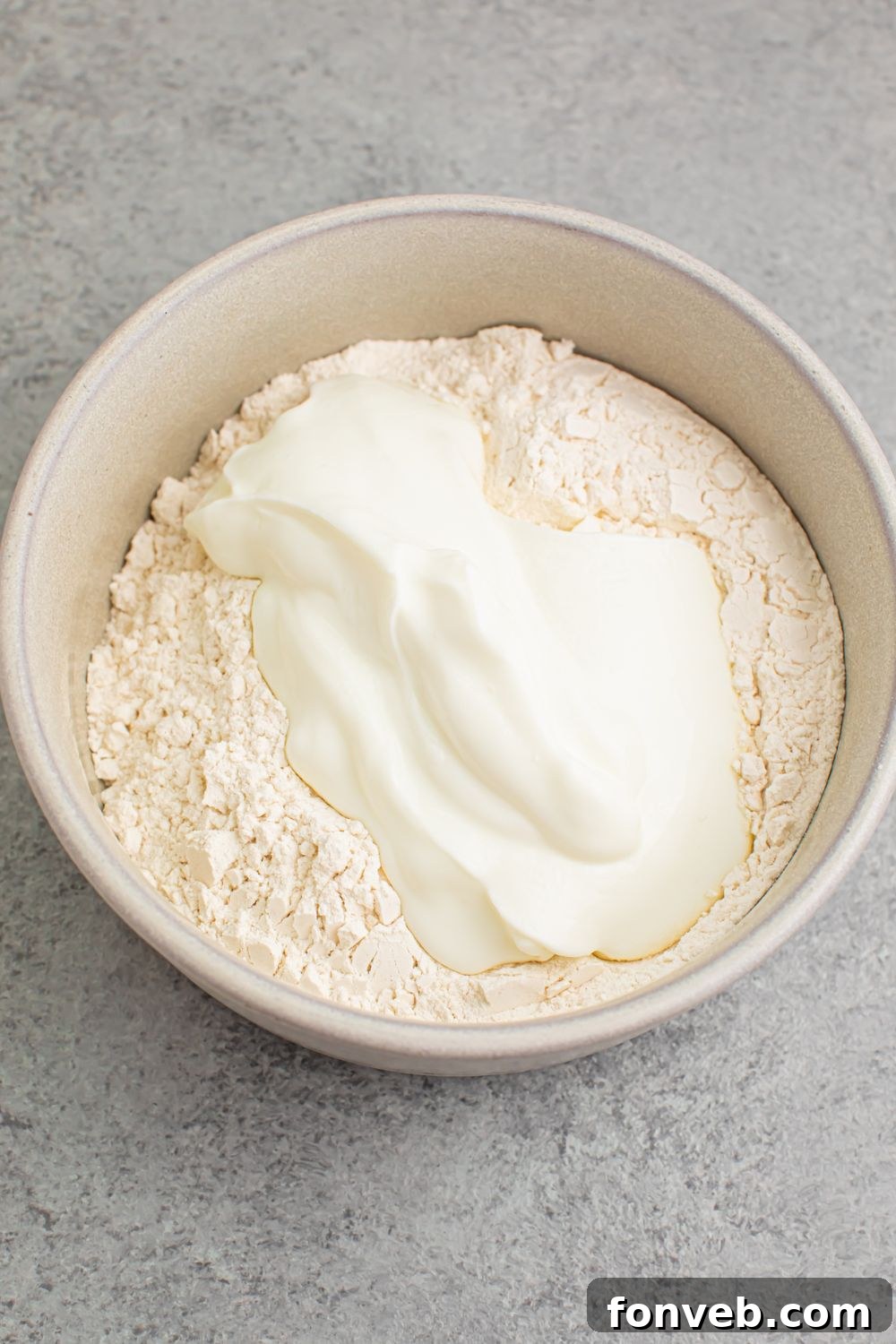 A white bowl on a wooden table, containing the initial mixture of self-rising flour and Greek yogurt, showing the start of the dough formation for 2-Ingredient Dinner Rolls.