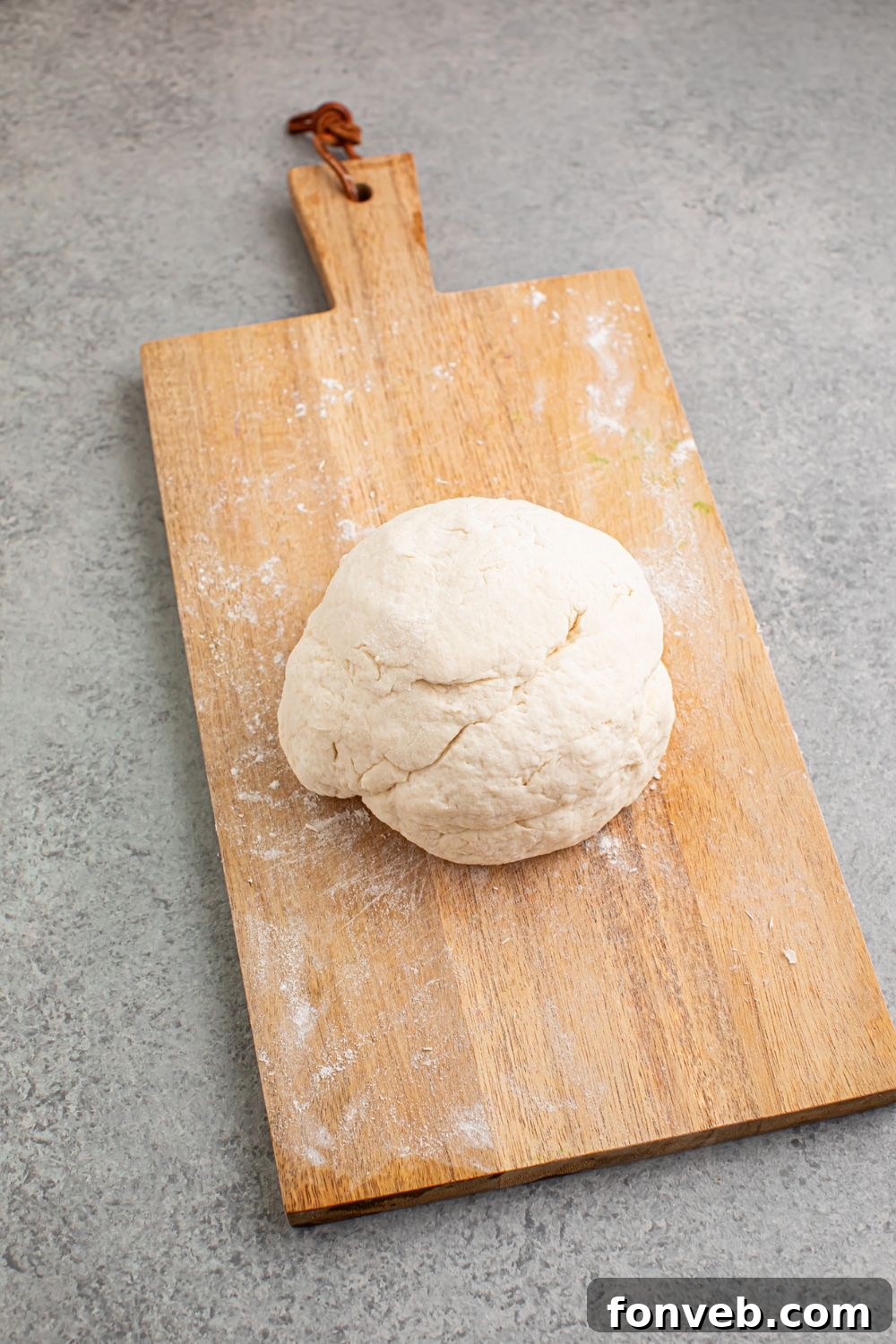 A large, soft dough ball, formed from self-rising flour and Greek yogurt, resting on a lightly floured cutting board, ready to be shaped into individual rolls.