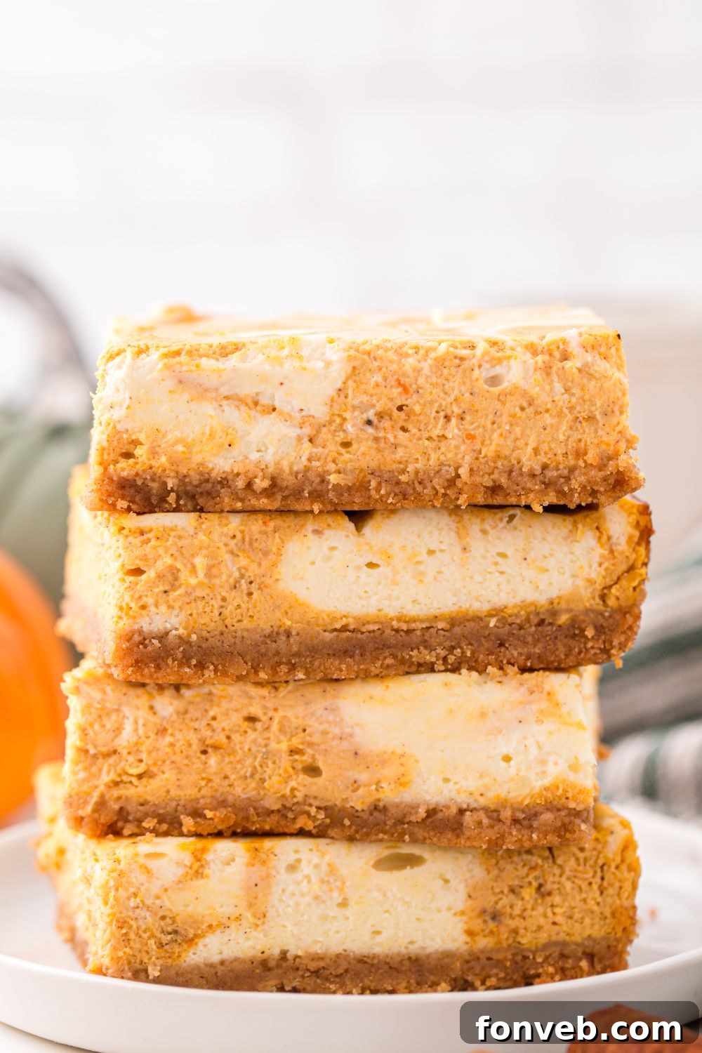Swirled Pumpkin Cheesecake Bars stacked on table with a towel and pumpkin behind it on table