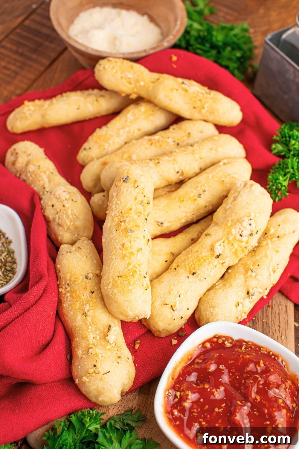 2-Ingredient Buttery Garlic Parmesan Breadsticks 13 red towel on table with 2 Ingredient Garlic Parmesan Breadsticks scattered on table with some dipping sauce in a bowl to the side