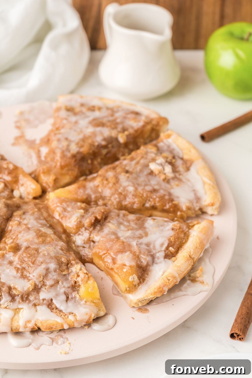 A dynamic side view of an Apple Pie Pizza on a wooden table, with cinnamon sticks and a vibrant green apple providing an appealing backdrop.