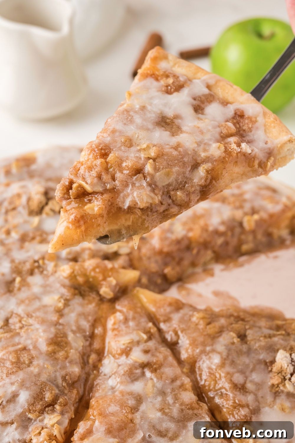 A slice of golden-brown Apple Pie Pizza is being lifted from a larger pizza on a wooden table, showcasing its rich apple filling and crumble topping.