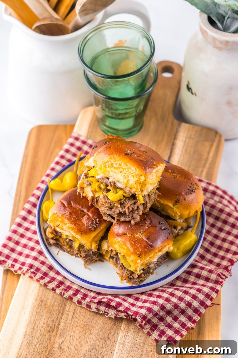 Deliciously Simple Slow Cooker Mississippi Pot Roast Sliders 15 Slow Cooker Mississippi Pot Roast Sliders stacked on a plate with a towel under it with some glasses behind it on table
