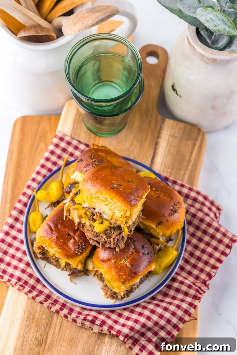 Deliciously Simple Slow Cooker Mississippi Pot Roast Sliders 21 Slow Cooker Mississippi Pot Roast Sliders stacked on a plate with some glasses and ceramic containers behind it on table
