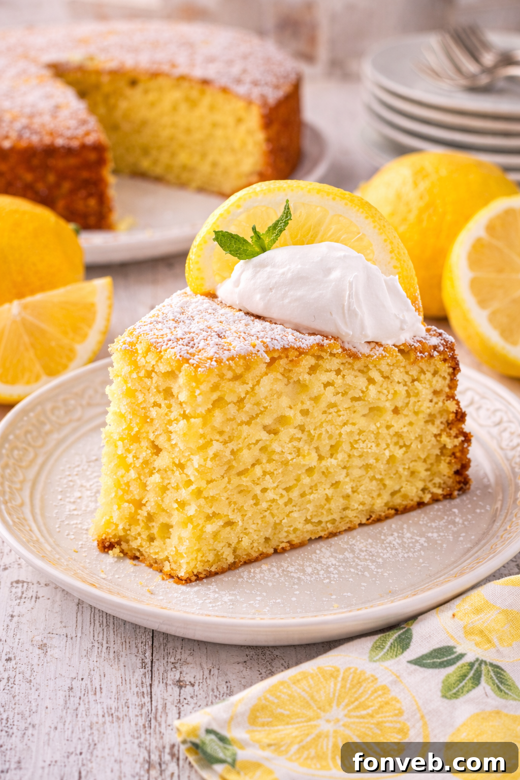 slice of Italian Lemon Ricotta Cake on a white plate with whole lemons behind it on table and the rest of the cake behind it