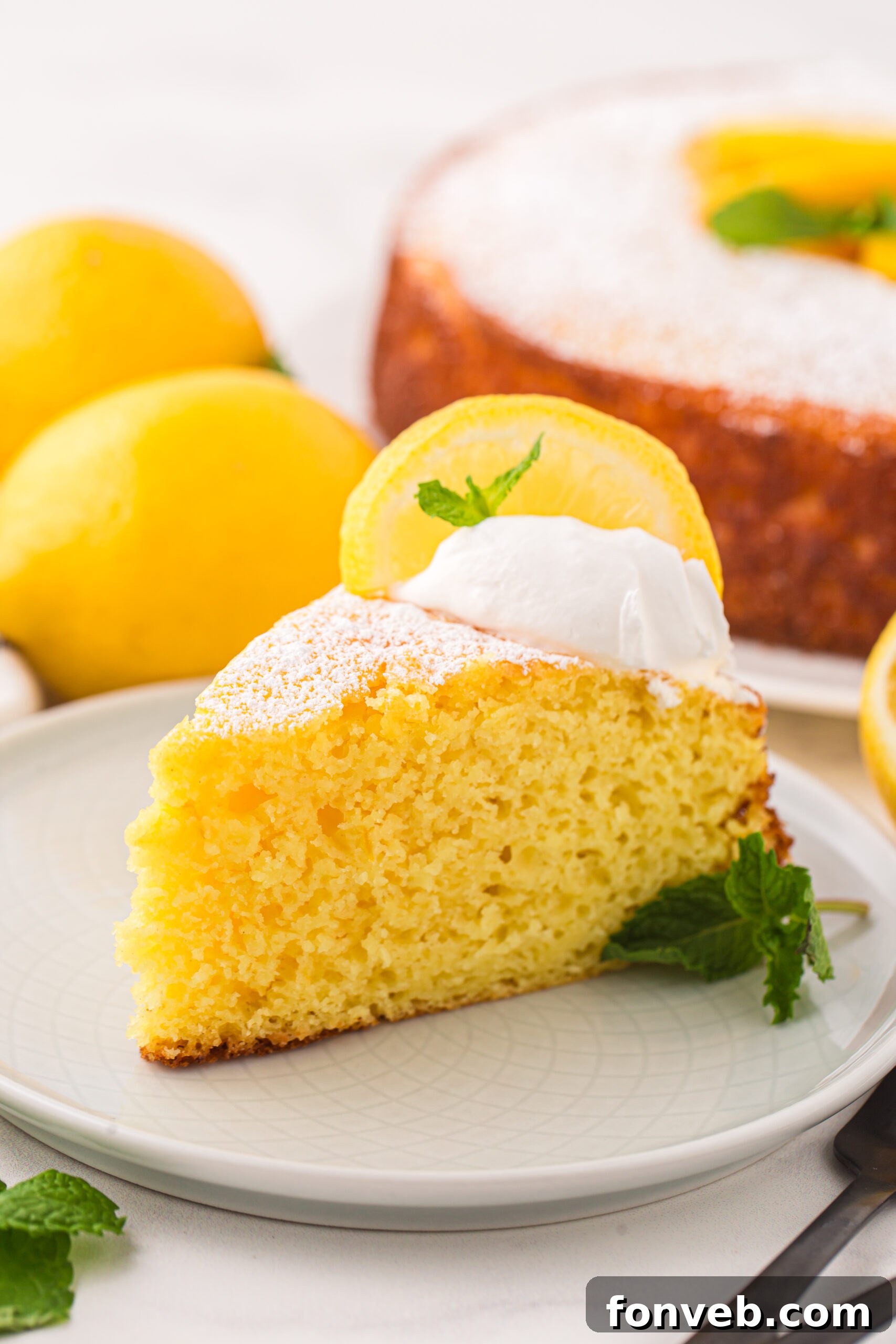 slice of Italian Lemon Ricotta Cake on a white plate with whole lemons behind it on table and the rest of the cake behind it