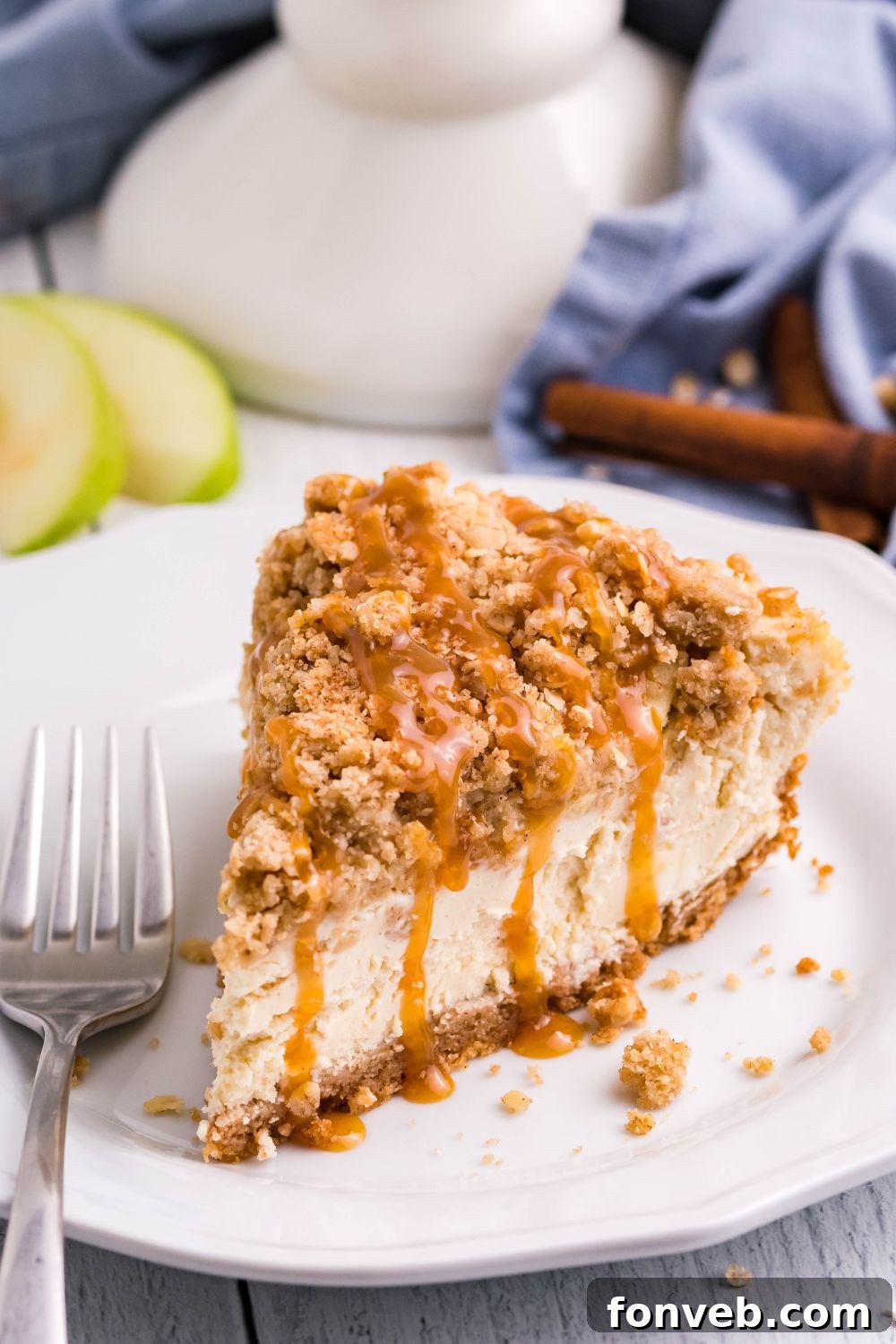 Decadent Caramel Apple Crumble Cheesecake 2 A perfectly sliced piece of Caramel Apple Crisp Cheesecake on a white plate, with whole apples and caramel sauce visible in the soft-focus background, highlighting its rich layers.