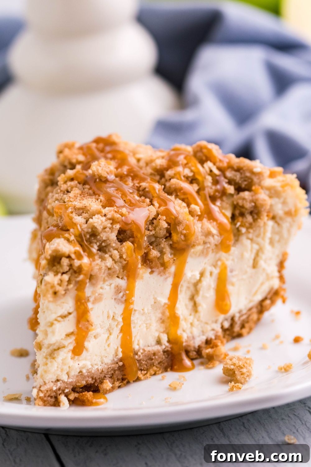Decadent Caramel Apple Crumble Cheesecake 11 A close-up of a slice of Caramel Apple Crisp Cheesecake on a plate, with a blue kitchen towel visible in the background, adding a touch of domestic warmth.
