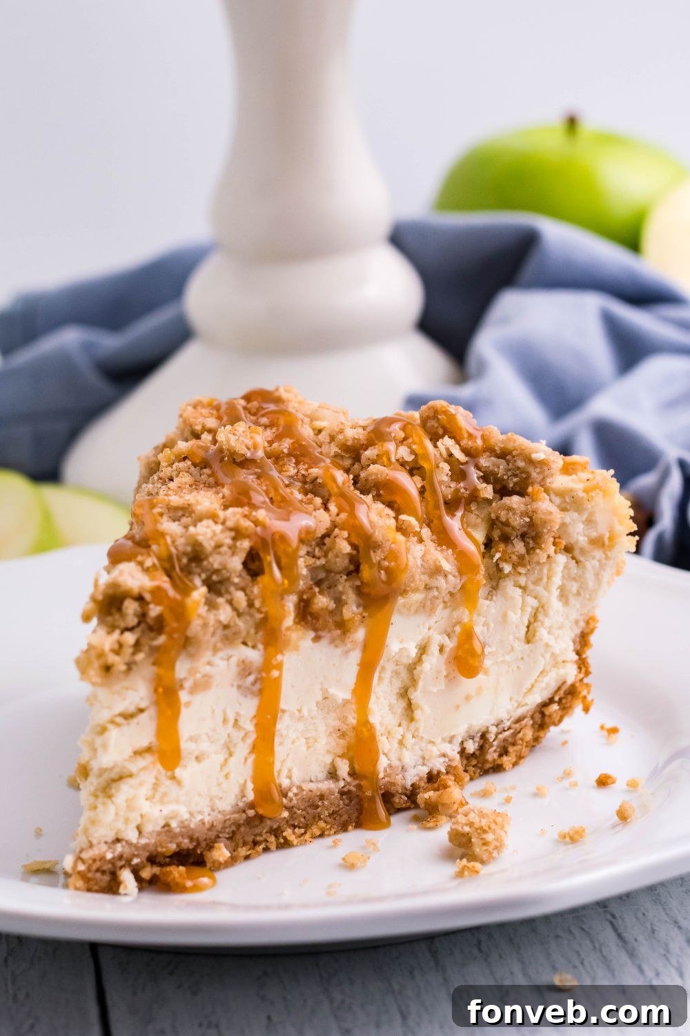 Decadent Caramel Apple Crumble Cheesecake 8 A slice of Caramel Apple Crisp Cheesecake on a plate with a pie plate, napkin, and green apples arranged in the background on a table, creating a rustic and appealing dessert tableau.