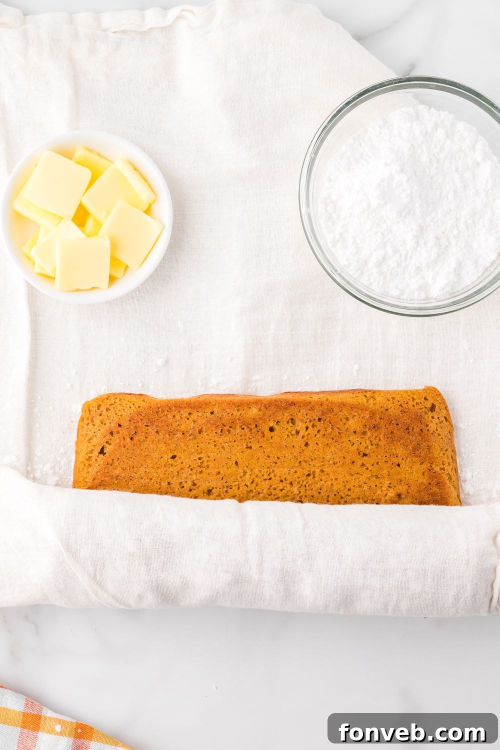 Classic Spiced Pumpkin Roll 12 rolling up a Pumpkin Cake Roll with parchment paper. To the side are two small bowls one with sliced butter the other with powdered sugar