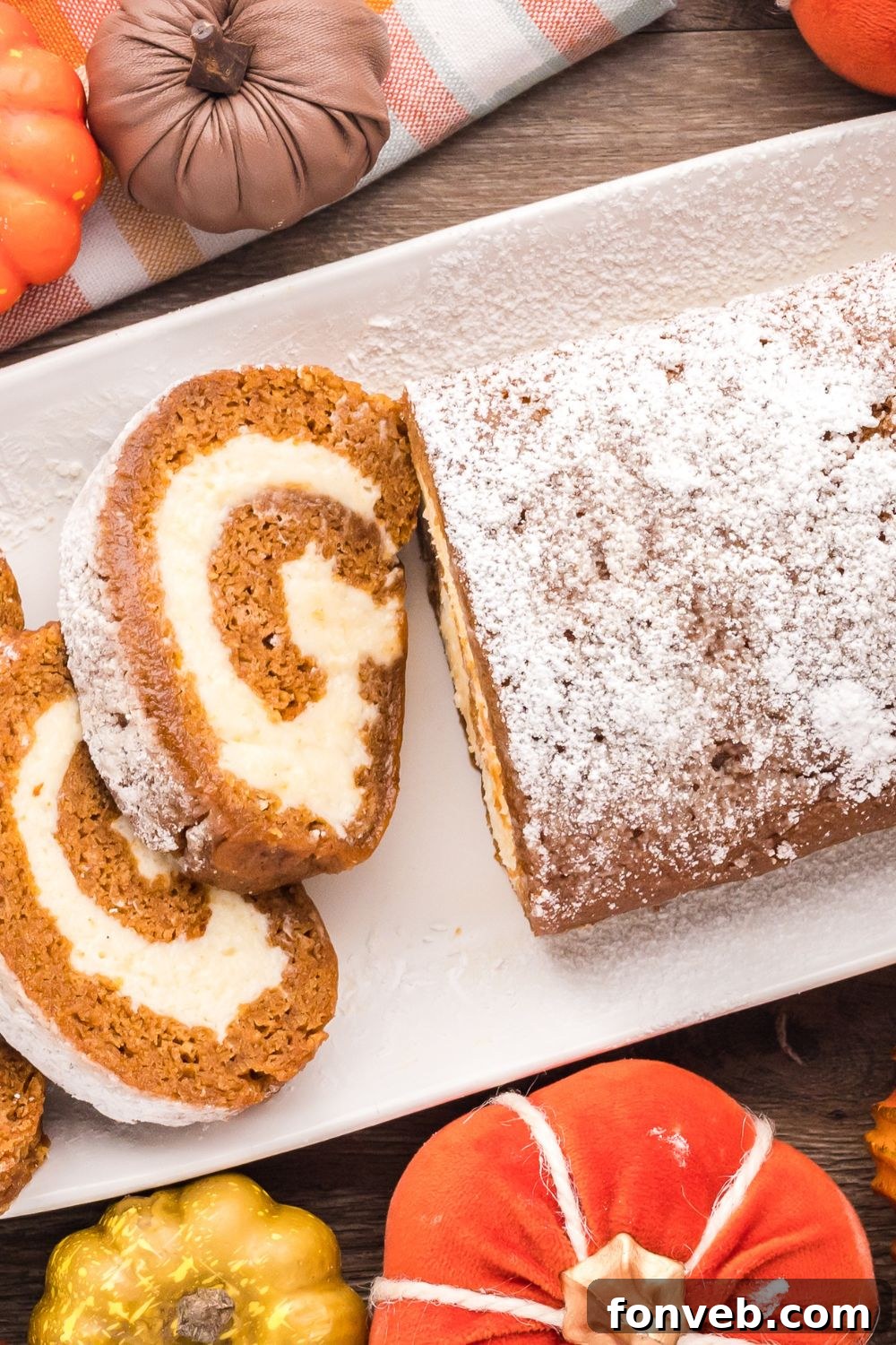 Classic Spiced Pumpkin Roll 21 Pumpkin Cake Roll on a long white platter, with part of it whole and the other part sliced. On the wooden table around the platter are mini pumpkins for a rustic fall display