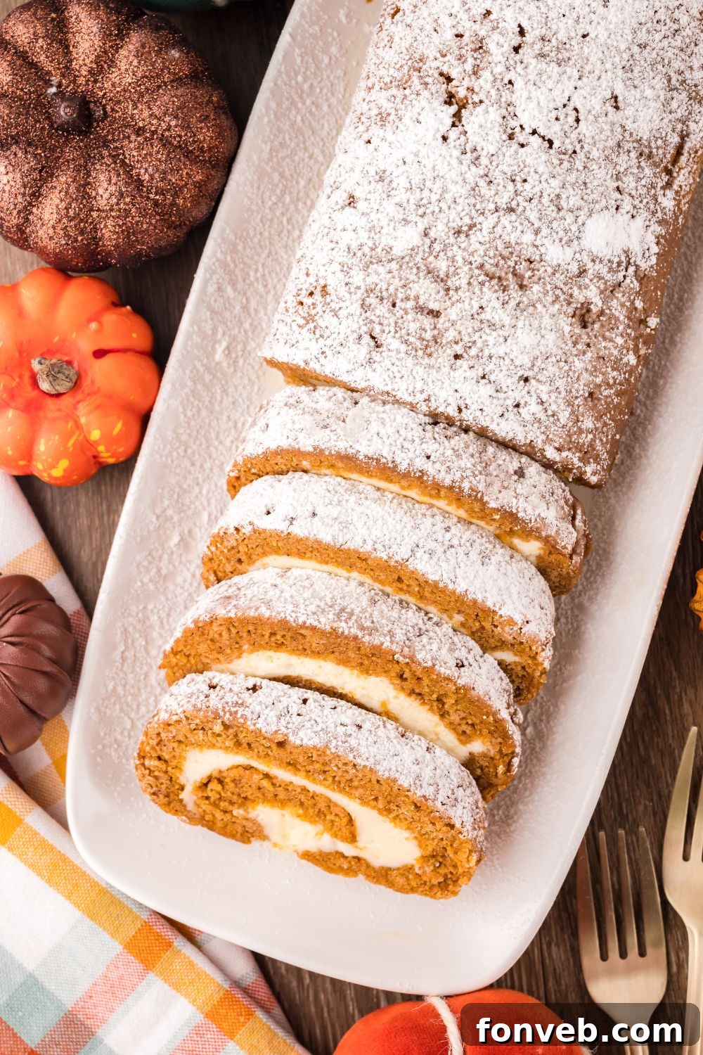 Classic Spiced Pumpkin Roll 23 overhead look down on a wooden table with fake mini pumpkins and a long Pumpkin Cake Roll on a white platter. Part of the roll is sliced into wedges