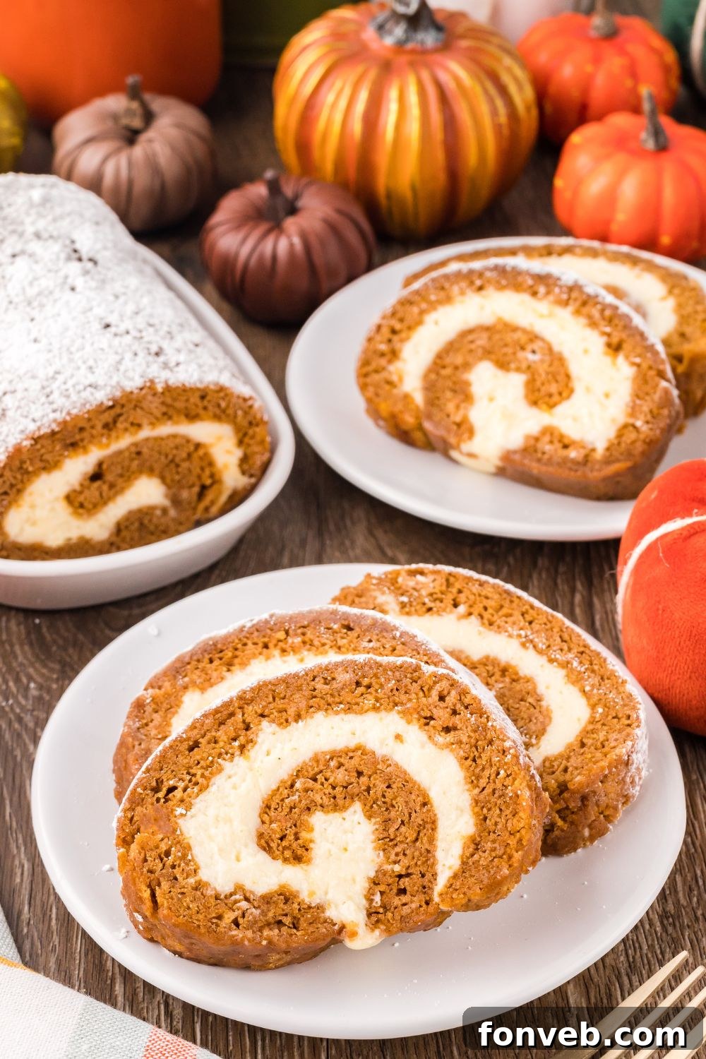 Classic Spiced Pumpkin Roll 25 Pumpkin Cake Roll placed on plates, with a small platter to side with more of the pumpkin roll. Mini pumpkins scattered on table for a Thanksgiving fall look