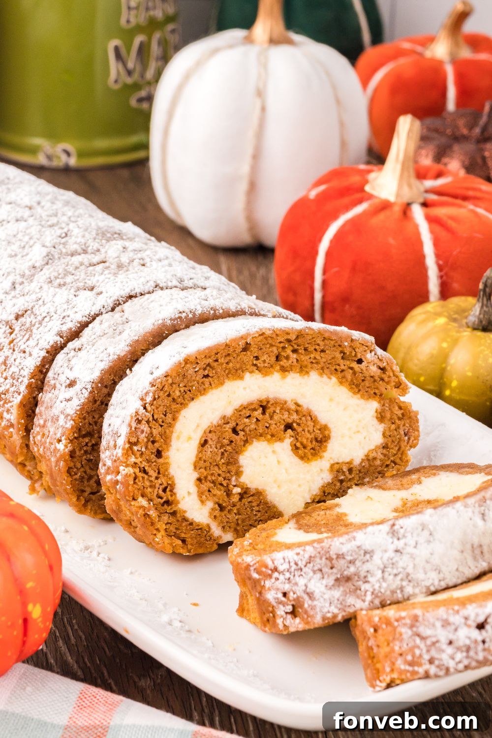 Classic Spiced Pumpkin Roll 28 Pumpkin Cake Roll placed on table sitting on a white platter, with a portion of the pumpkin cake sliced. Then you have a mix of faux pumpkins scattered on table for a rustic fall look