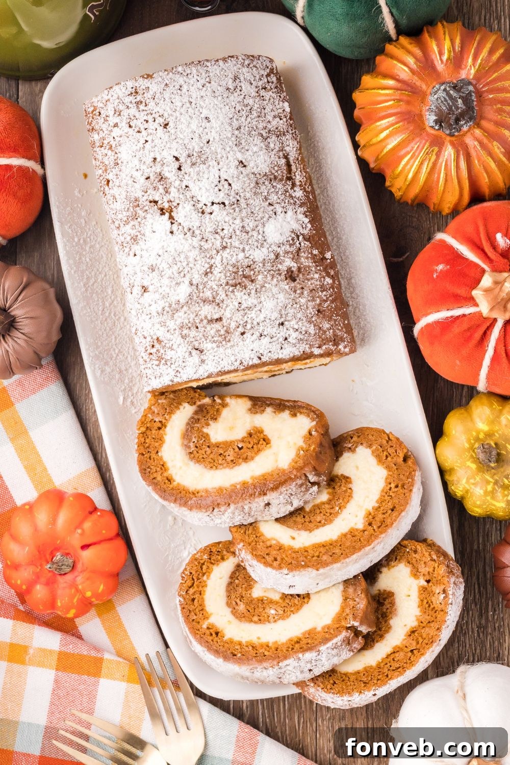 Classic Spiced Pumpkin Roll 5 Pumpkin Cake Roll on a long white platter, with half of the pumpkin roll sliced. Faux pumpkins around the table with a white and brown striped towel