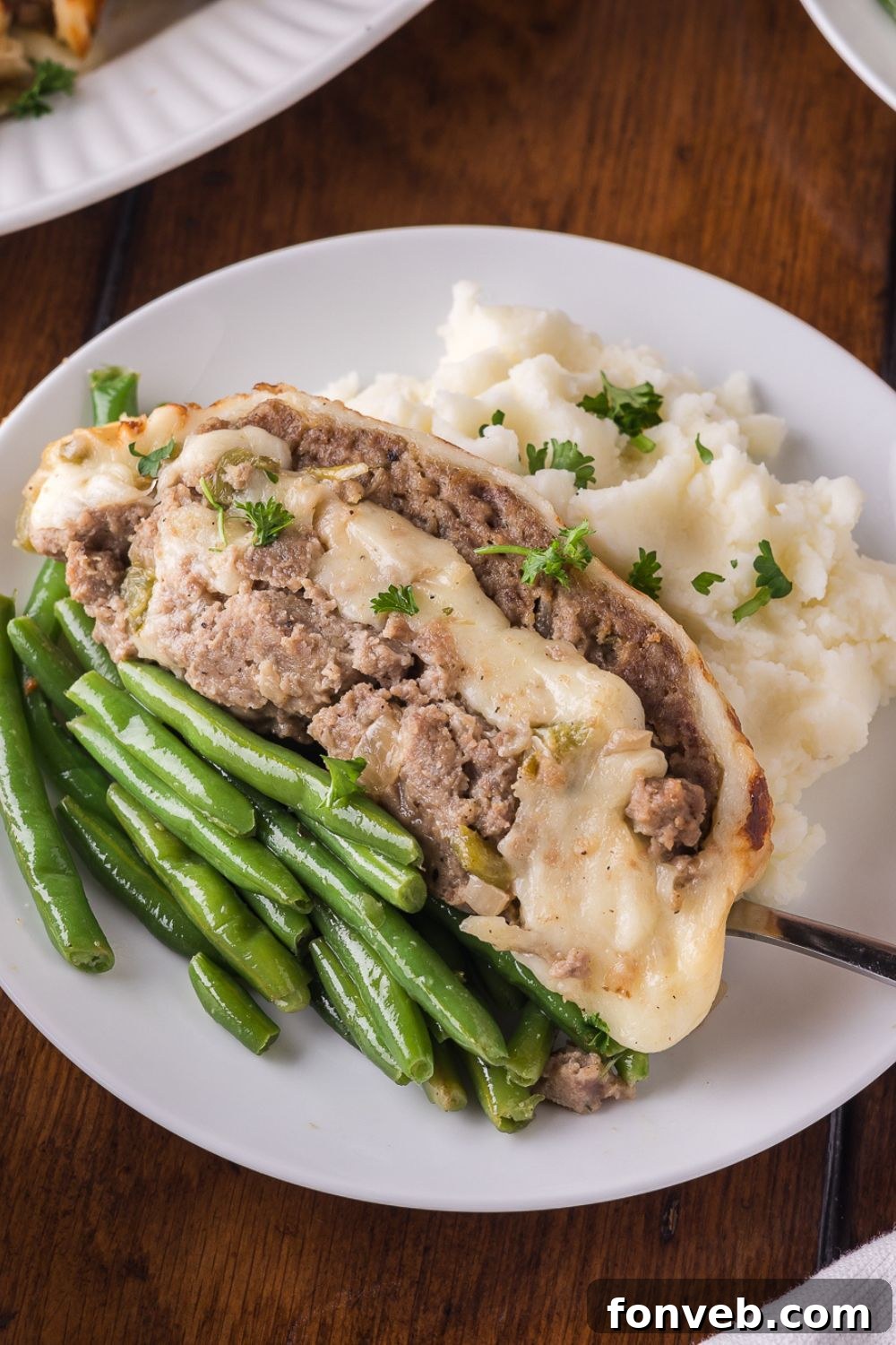 wooden table with a white plate sitting on it, that has green beans, philly cheesesteak meatloaf, and a spoonful of mashed potatoes
