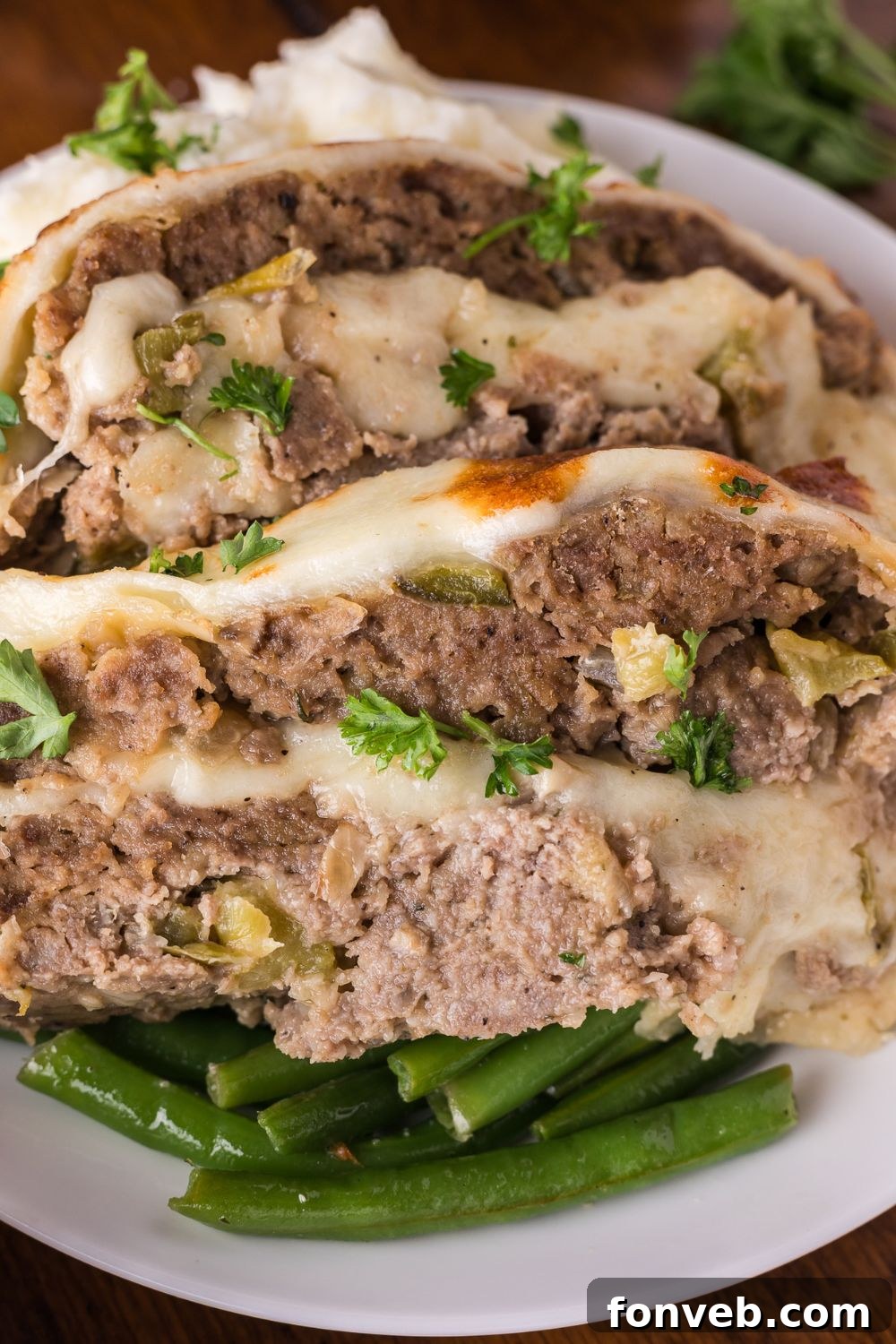 white plate sitting on table with a bed of green beans, and on top thick slices of Stuffed Philly Cheesesteak Meatloaf topped with fresh parsley
